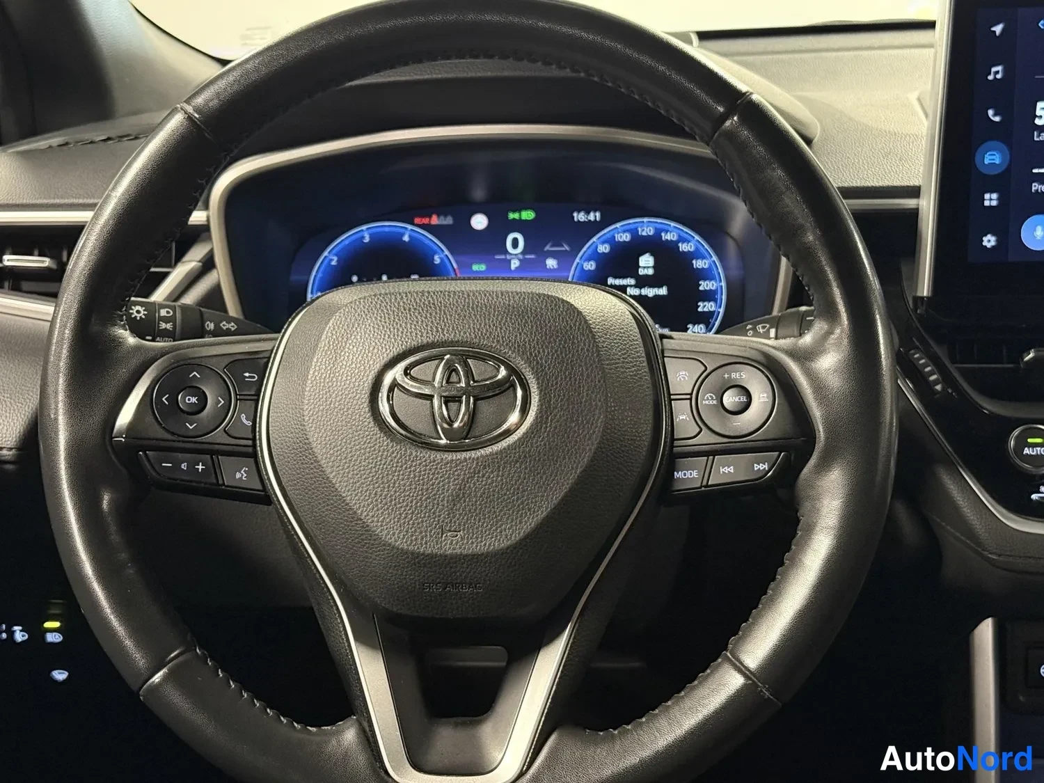Sivulta: Ohjauspyörän lähikuva Toyota-logolla ja monitoimipainikkeilla (mm. OK, puhe, äänenvoimakkuus ja 2-asteinen muisti), taustalla digitaalinen mittaristo.