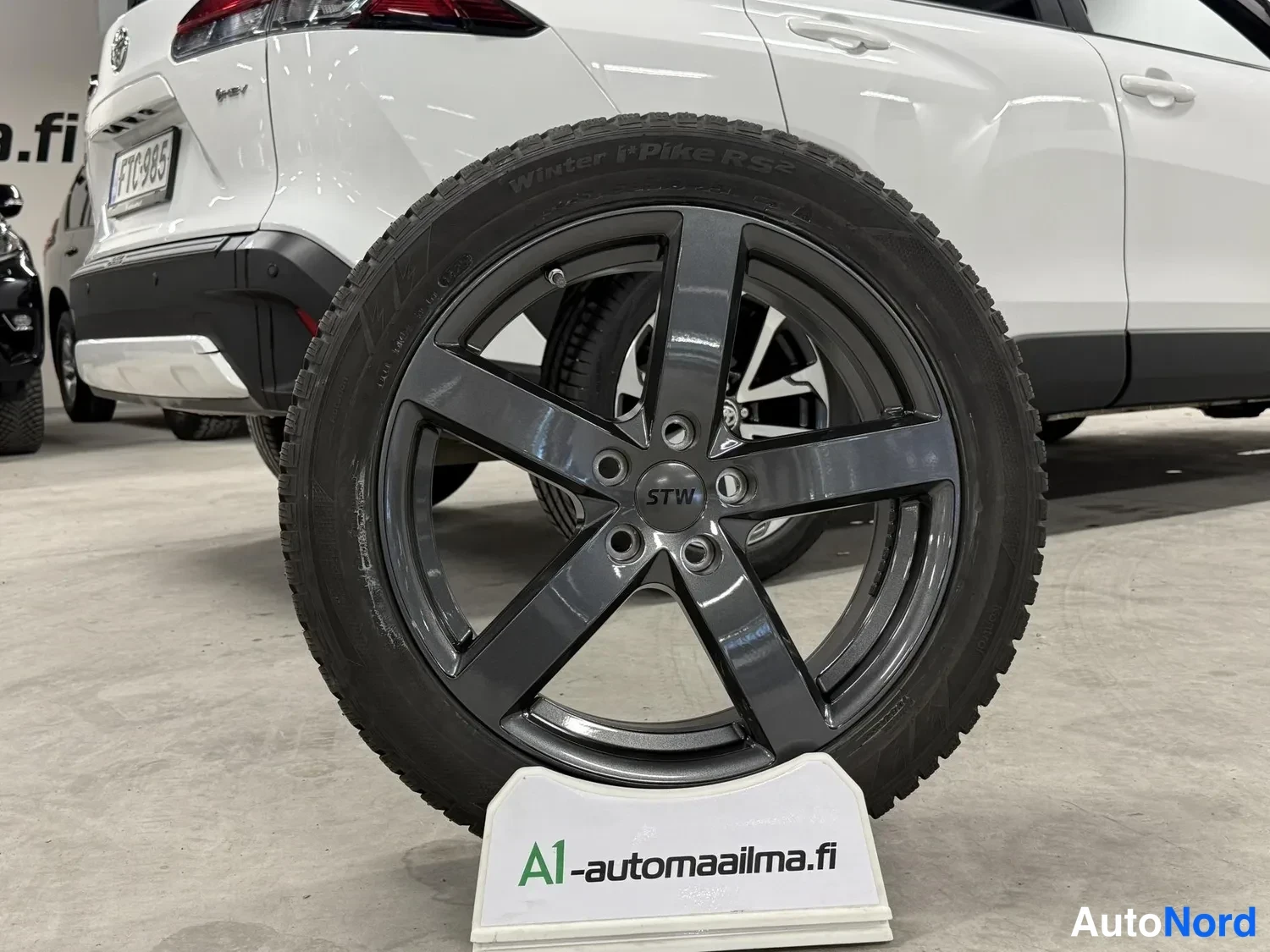 Takaviisto: Talvirengas Winter Spike RS2 tummassa viisipuomaisessa STW-vanteessa jalustalla, taustalla valkoinen auton perä.