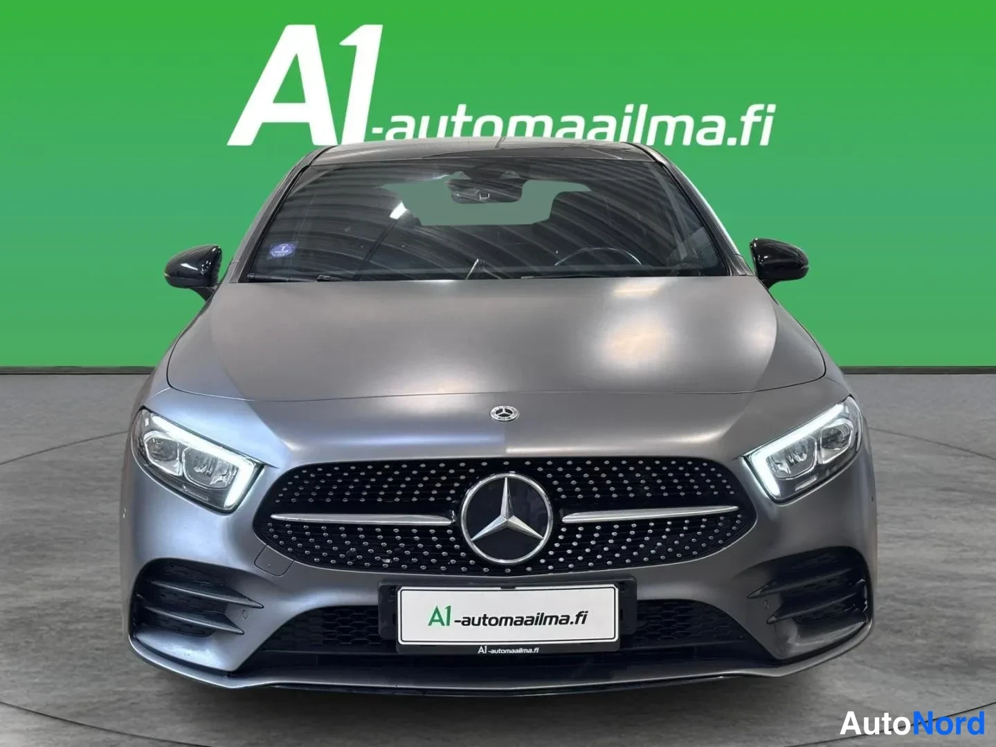 Edestä: Mercedes-Benz A-sarjan sedanin etunäkymä, harmaa kori, tähtikoriin säböpositio, A1-automaailma-taustalla.