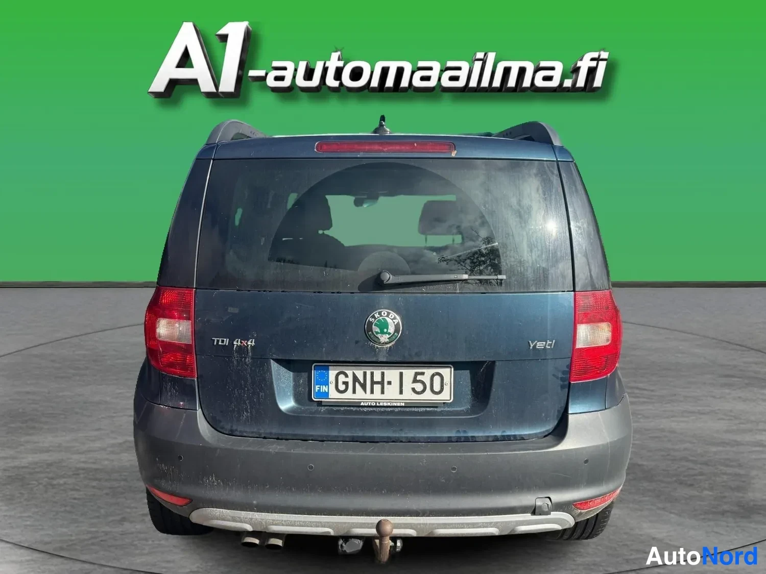 Takaa: Auton takaosa suoraan takaa: tummansininen Skoda Yeti, rekisteritunnus, kattokaiteet, vetokoukku ja kaksoispakoputki näkyvissä vihreällä taustalla.