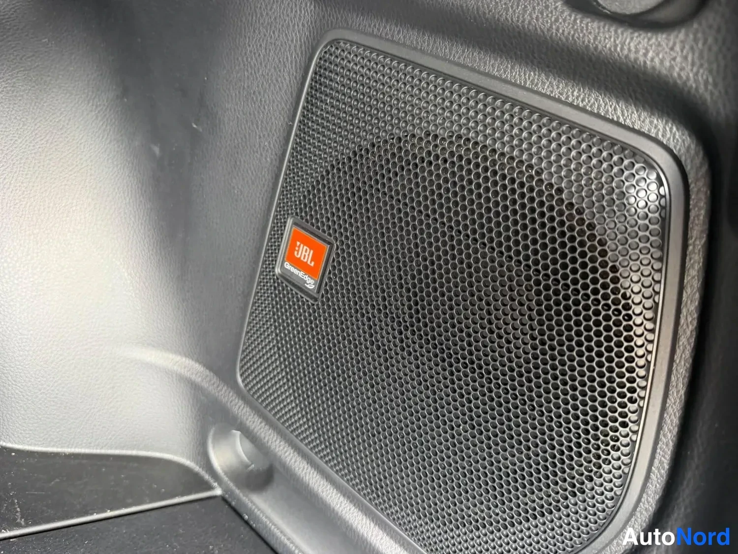 Muu: Sisäkolmikulman kuva autoklin alla näkyvästä kaiuttimesta: musta verkko ja oranssi JBL GreenEdge -merkki.