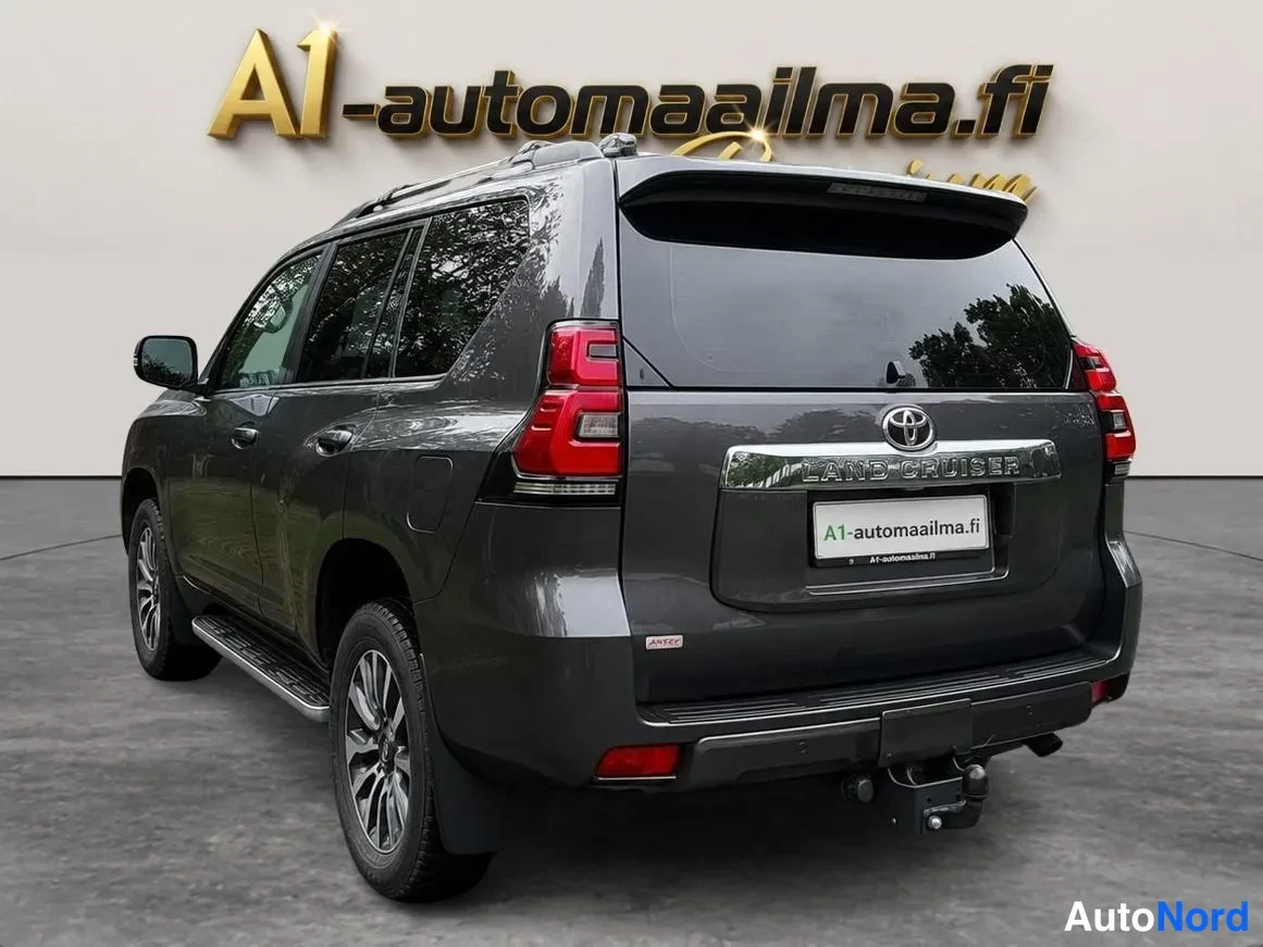 Takaviisto: Tummanharmaa Toyota Land Cruiser -henkinen maastopyörä takakolmannesvyöhykkeestä, perä oikealla; takavalot, LAND CRUISER -teksti ja kattokaiteet näkyvissä.