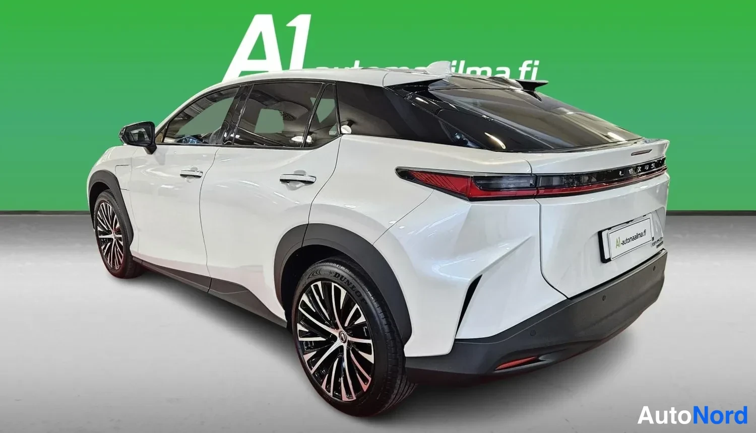 Takaviisto: Ajetta valkoinen Lexus RZ -maasturi takaa sivulta kuvattuna, näkyvät takavalot, rekisterikilpi ja monihaaraiset kevytmetallivanteet vihreää taustaa vasten.
