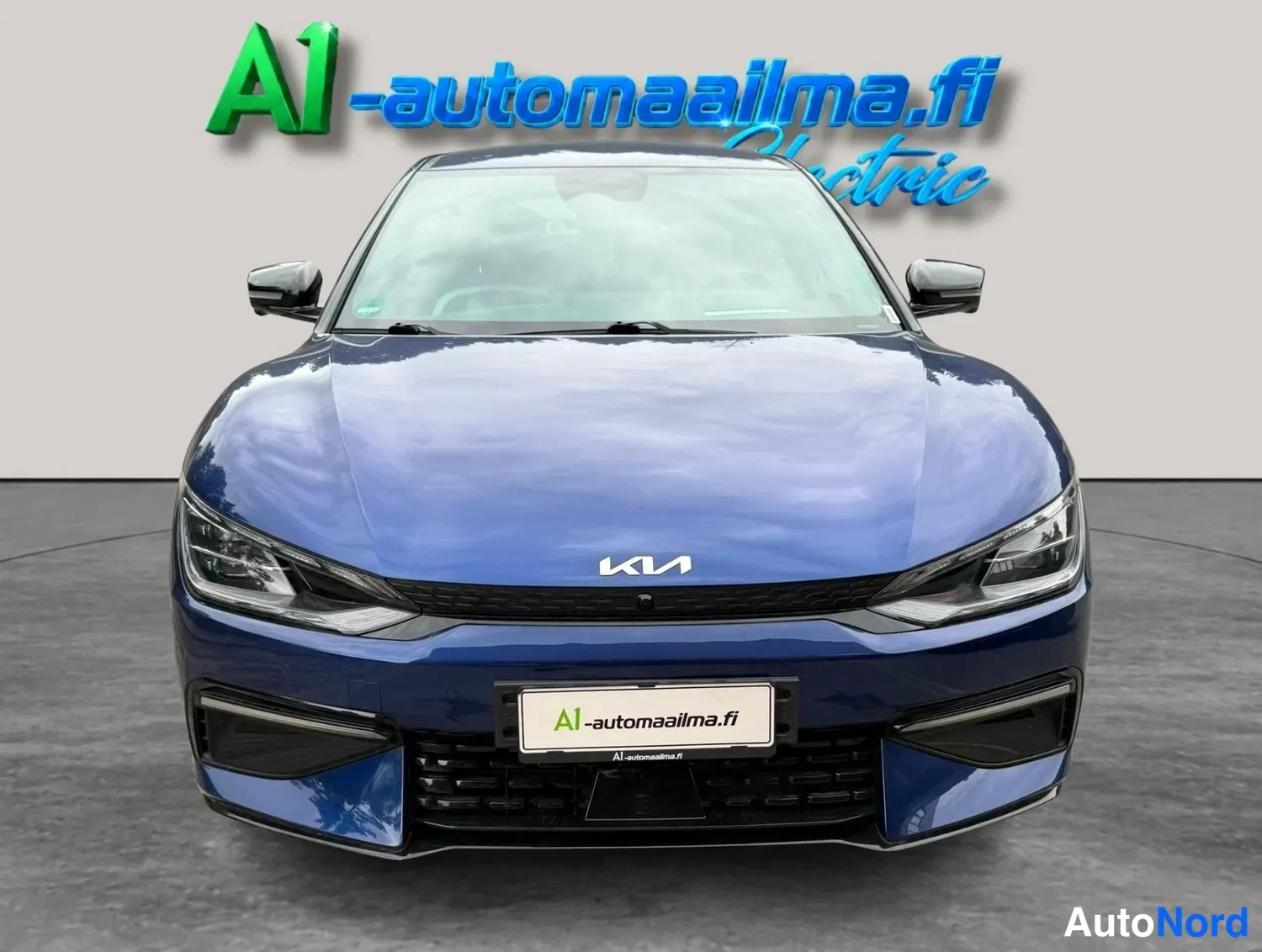 Edestä: Sininen Kia EV6 suoraan edestä, KIAsymboli konepellissä ja A1-automaailma.fi-kyltti etupuskurissa.