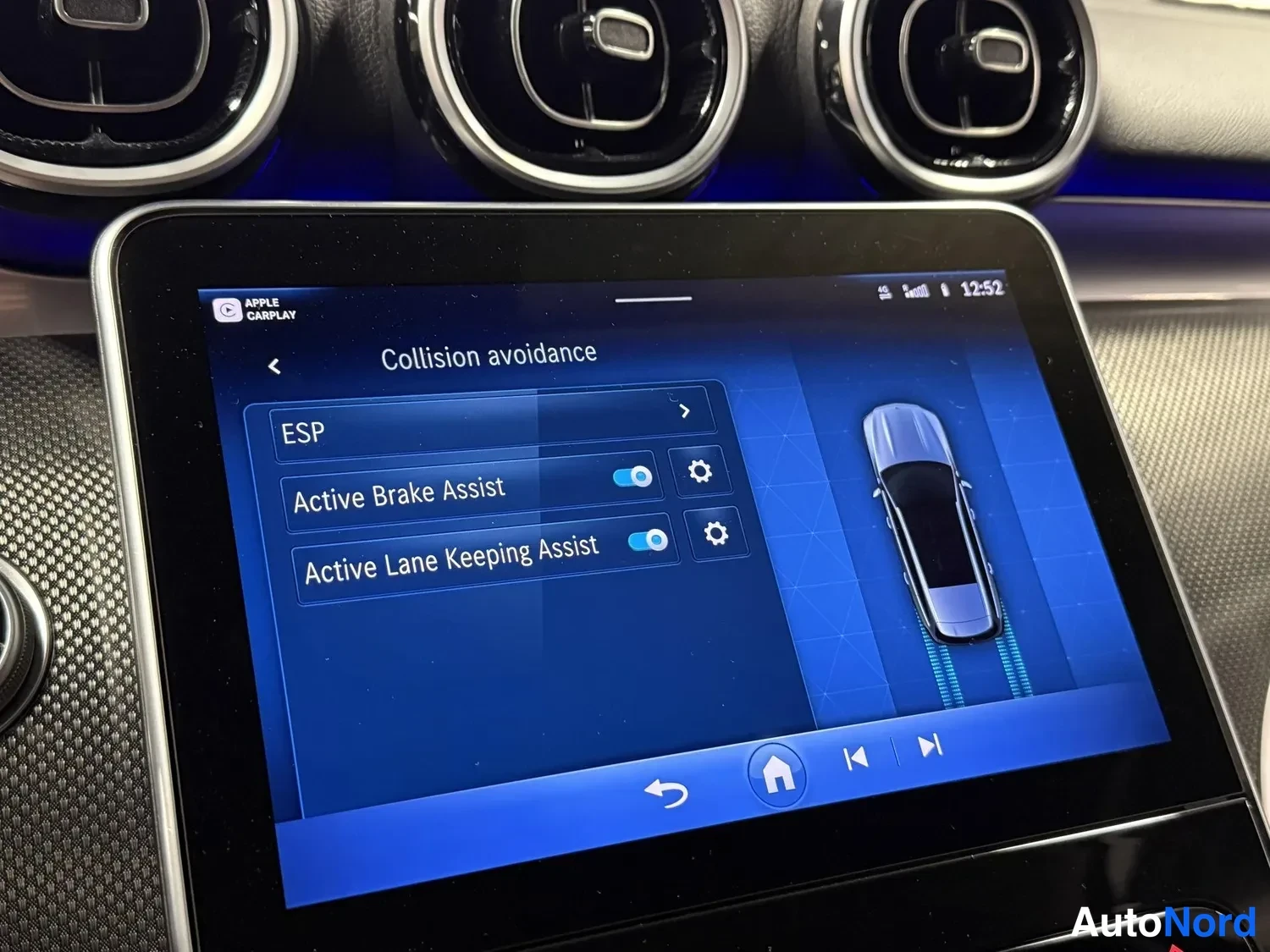 Yksityiskohta: Kojelaudan kosketusnäyttö, jossa näkyy Collision avoidance -valikko: ESP, Active Brake Assist ja Active Lane Keeping Assist päällä. Näytön yläkulmassa Apple Car