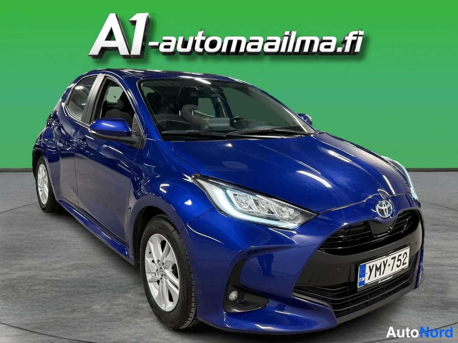 Etuviisto, nokka oikealle: Sininen Toyota Yaris -henkilöauto etu- ja sivusta kolmeneljäsosan kuvakulmasta, tunnus.