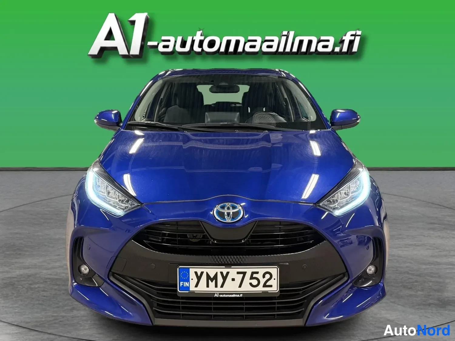 Edestä: Sininen Toyota Yaris -henkilöauto suoraan edestä kuvattuna, tunnus.