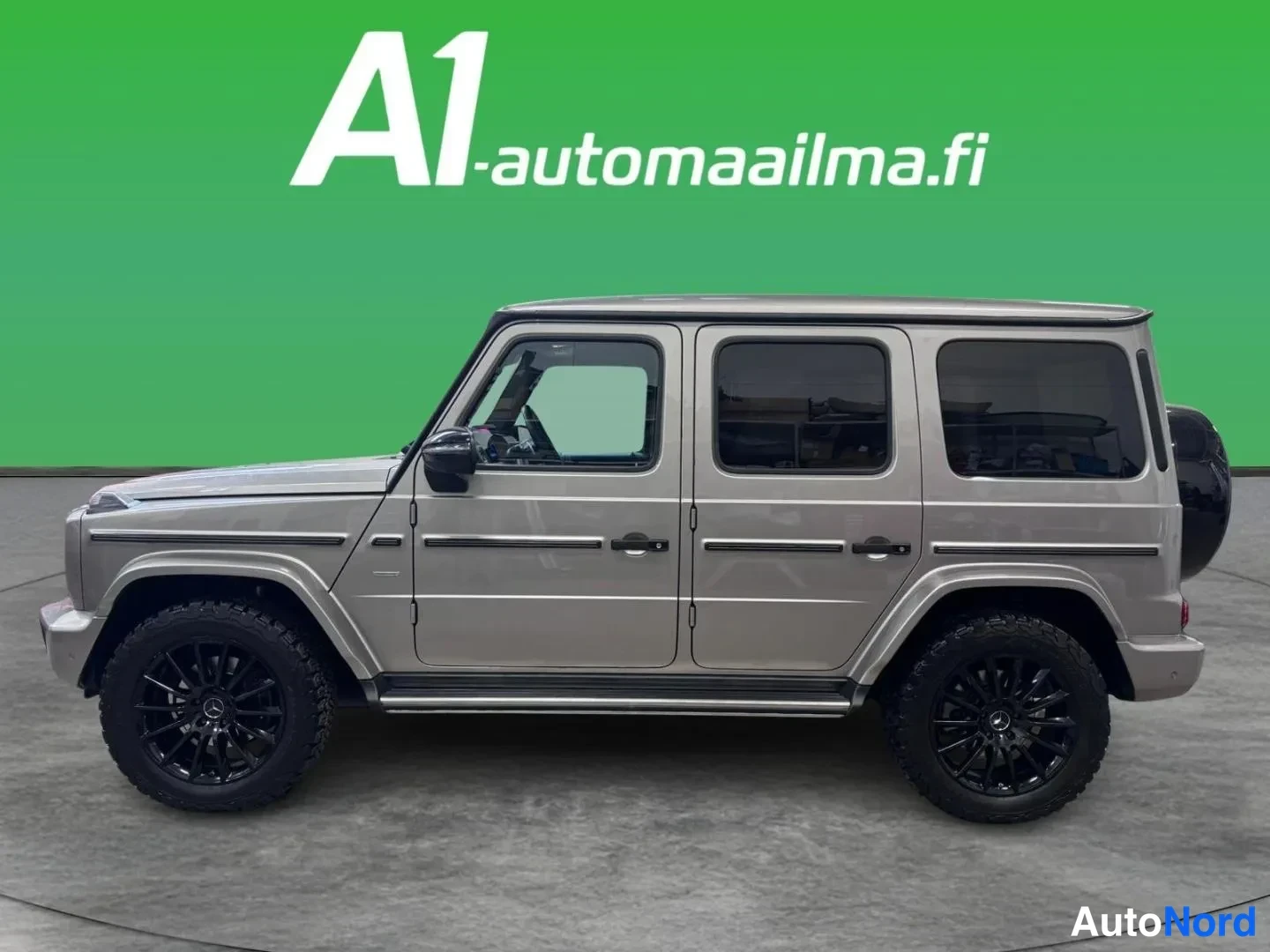 Sivulta: Mercedes-Benz G:n sivukuva autosalissa: harmaa beige koripelti, neljä ovea, mustat vanteet ja kattotelineen pohja näkyvät.