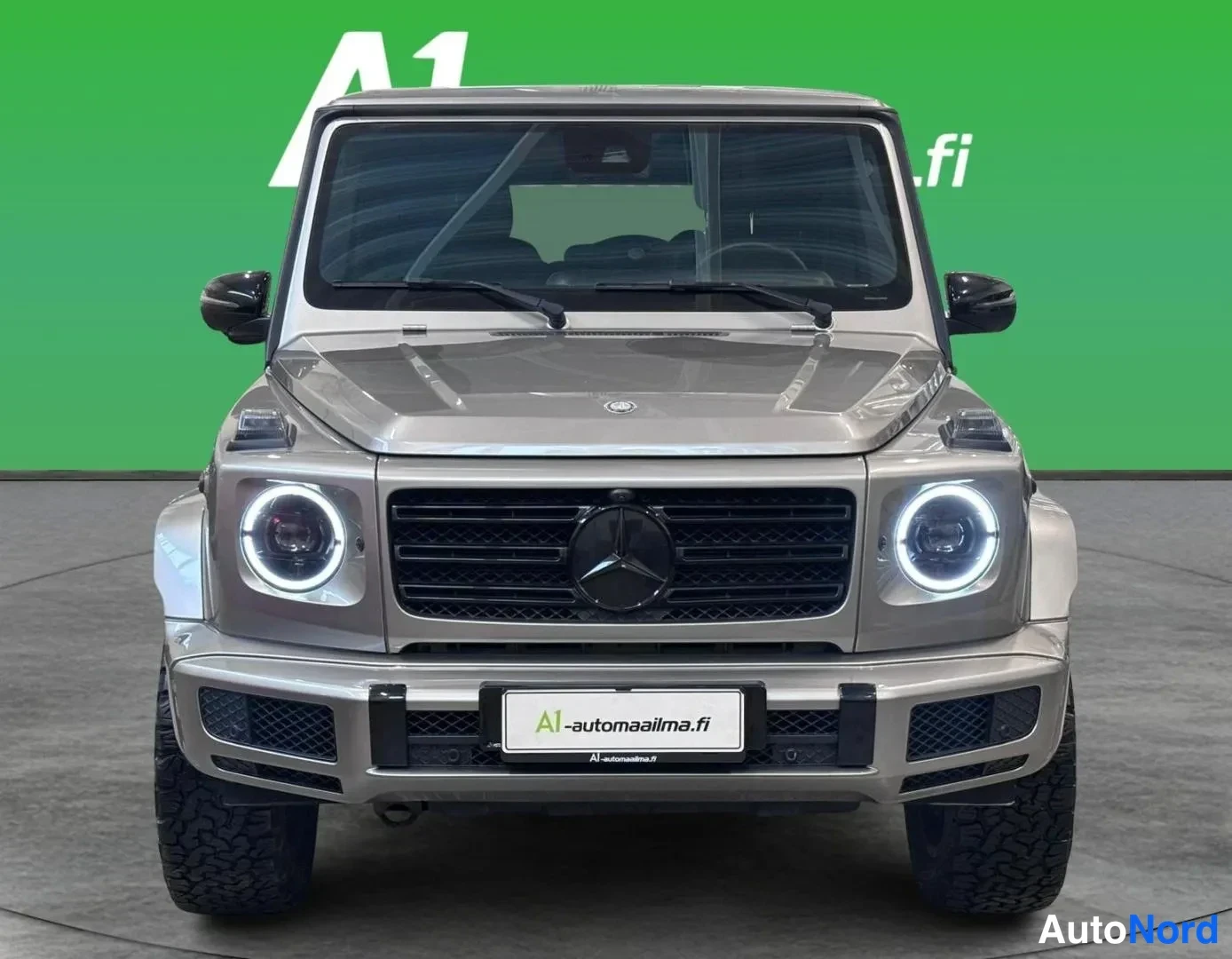 Edestä: Mercedes-Benz G:n suora etukuva autosalissa: harmaa beige konepelti, musta säleikkö Mercedes-tähdellä ja rengasmaiset LED-merkkivalot.