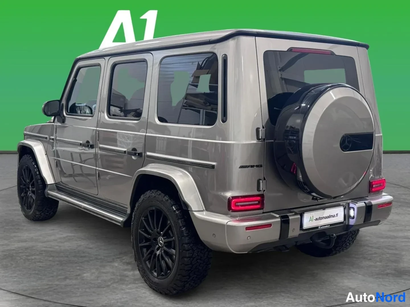 Takaviisto: Mercedes-Benz G -luokan takakulmanäkymä vasemmalta, harmaasävyinen maasturi varapyörällä takaluukussa ja mustilla vanteilla.