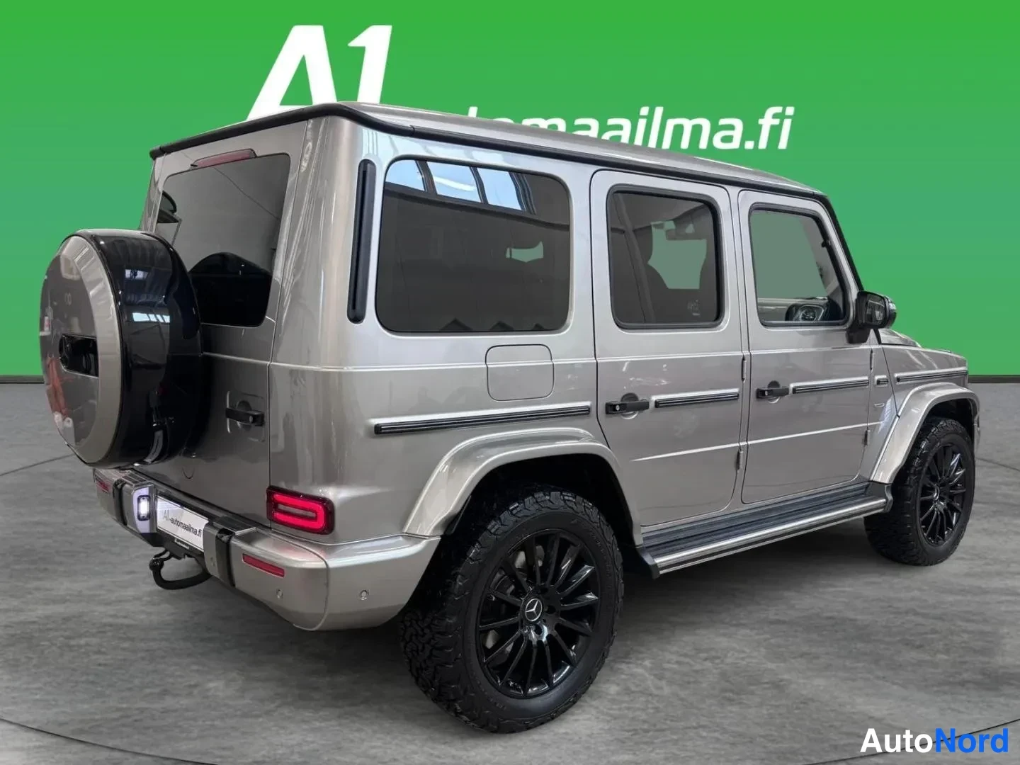 Takaviisto: Mercedes-Benz G -luokan takakulmanäkymä vasemmalta, harmaasävyinen maasturi mustilla vanteilla ja sivulistoilla.