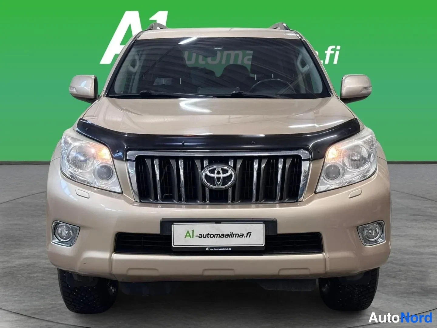 Edestä: Beige Toyota Land Cruiser suoraan edestäpäin, keulagridi, Toyota-merkki ja ajovalot näkyvät studiopohjalla.