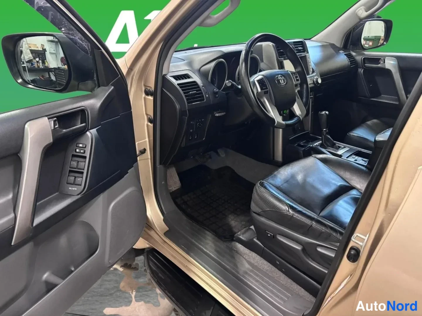 Muu: Avoimen etuoven sisäpuoli ja ohjaamo: Toyota-merkkinen ohjauspyörä, manuaalikkivalot ovessa, tummat nahkaistuimet ja tiedostolaatikon tumma matto lattialla.