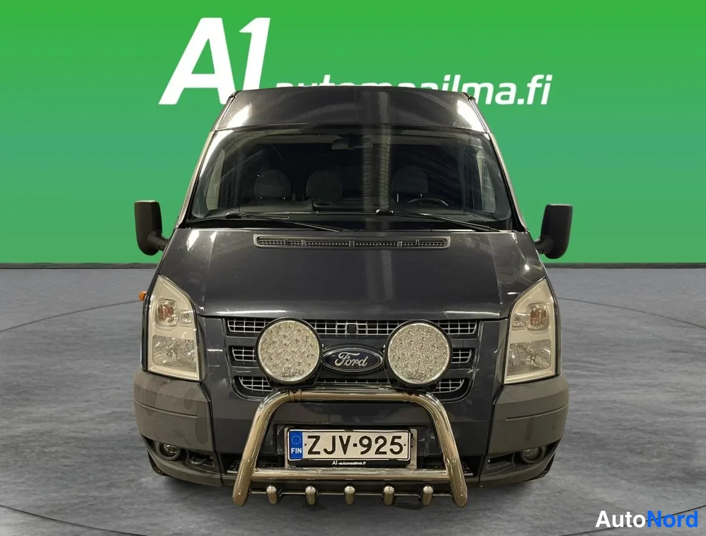 Edestä: Ford Transit -pakettiauto suoraan edestä, tummanharmaa kori, jäykkä törmäyssuoja ja kaksi LED-lisävaloa, rekisterikilpi.