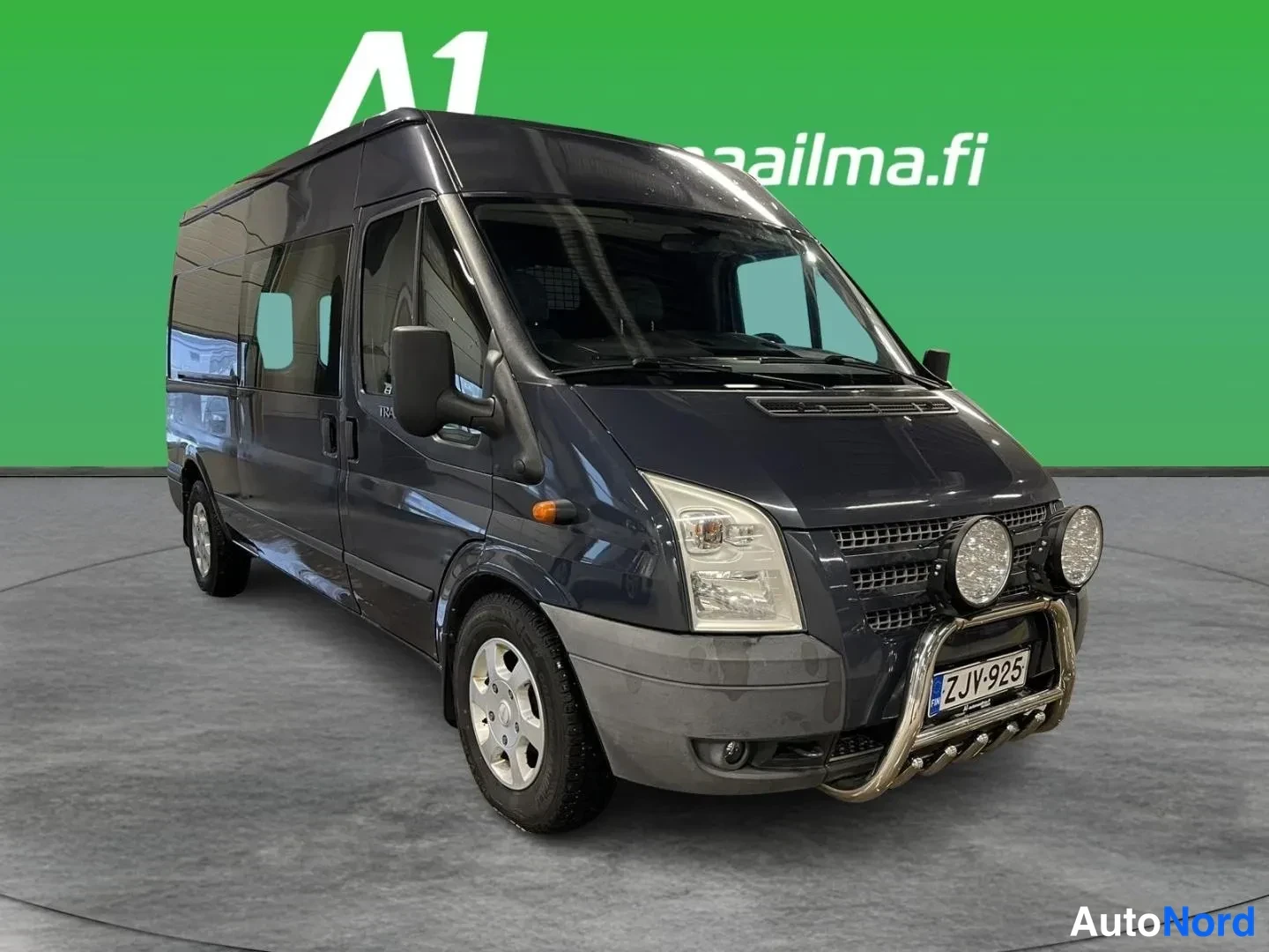 Etuviisto, nokka oikealle: Ford Transit -pakettiauto etuviistosta oikealta, tummanharmaa kori, sivuiltaan näkyvä korirakenne, etupuskurin osa ja kaksi lisävaloa, rekisterikilpi.