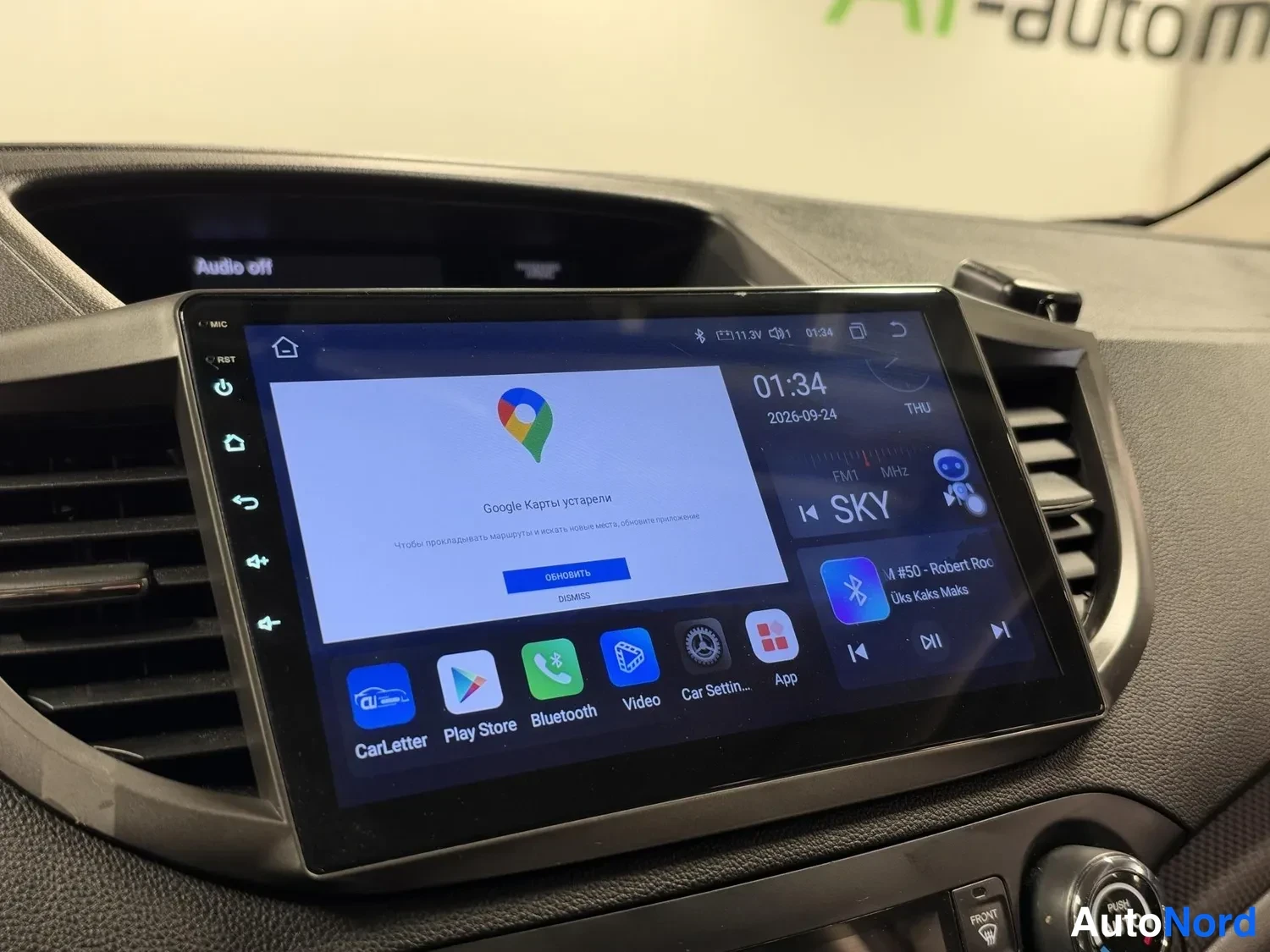 Muu: Kojelaudan Android-pohjainen kosketusnäyttö, jossa Google Maps -sovellus, kellonaika 01:34 ja radioasema SKY
