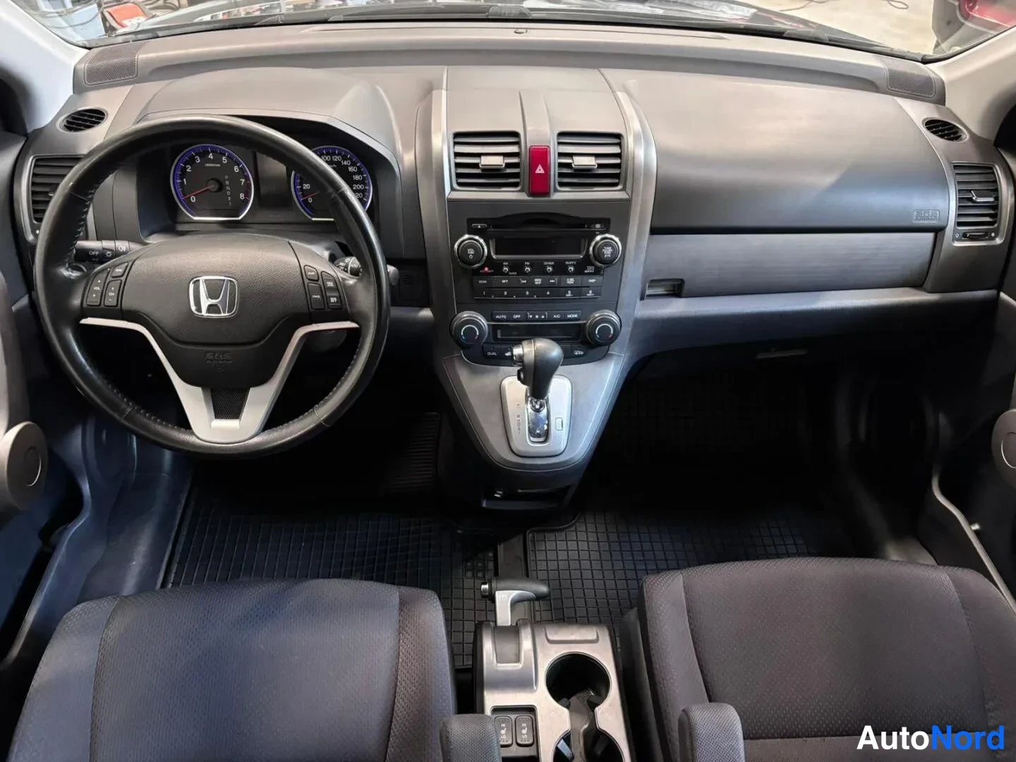 Muu: Honda CR-V:n ohjaamo: tumma kojelauta, Honda-ohjauspyörä, analogiset mittarit, automaattivaihteen valitsin ja radiopaneeli; etupenkit ja lattiamatat näkyvät.