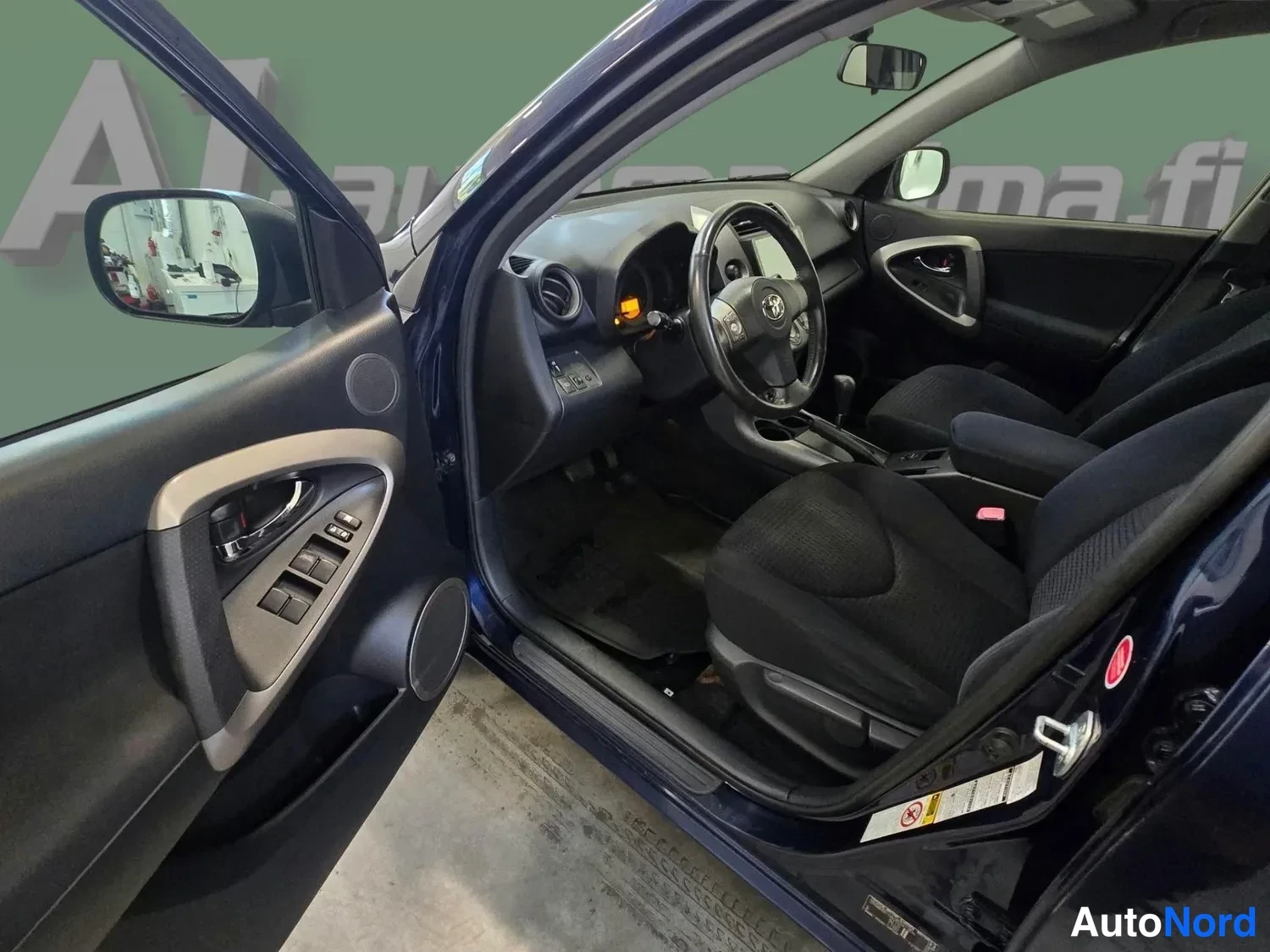 Muu: Toyota RAV4:n etusisätilat kuljettajan oven auki: rattitilan ohjauspyörä, automaattivaihteen valitsin, tummat kangasistuimet ja ovinapeikon säätimet.