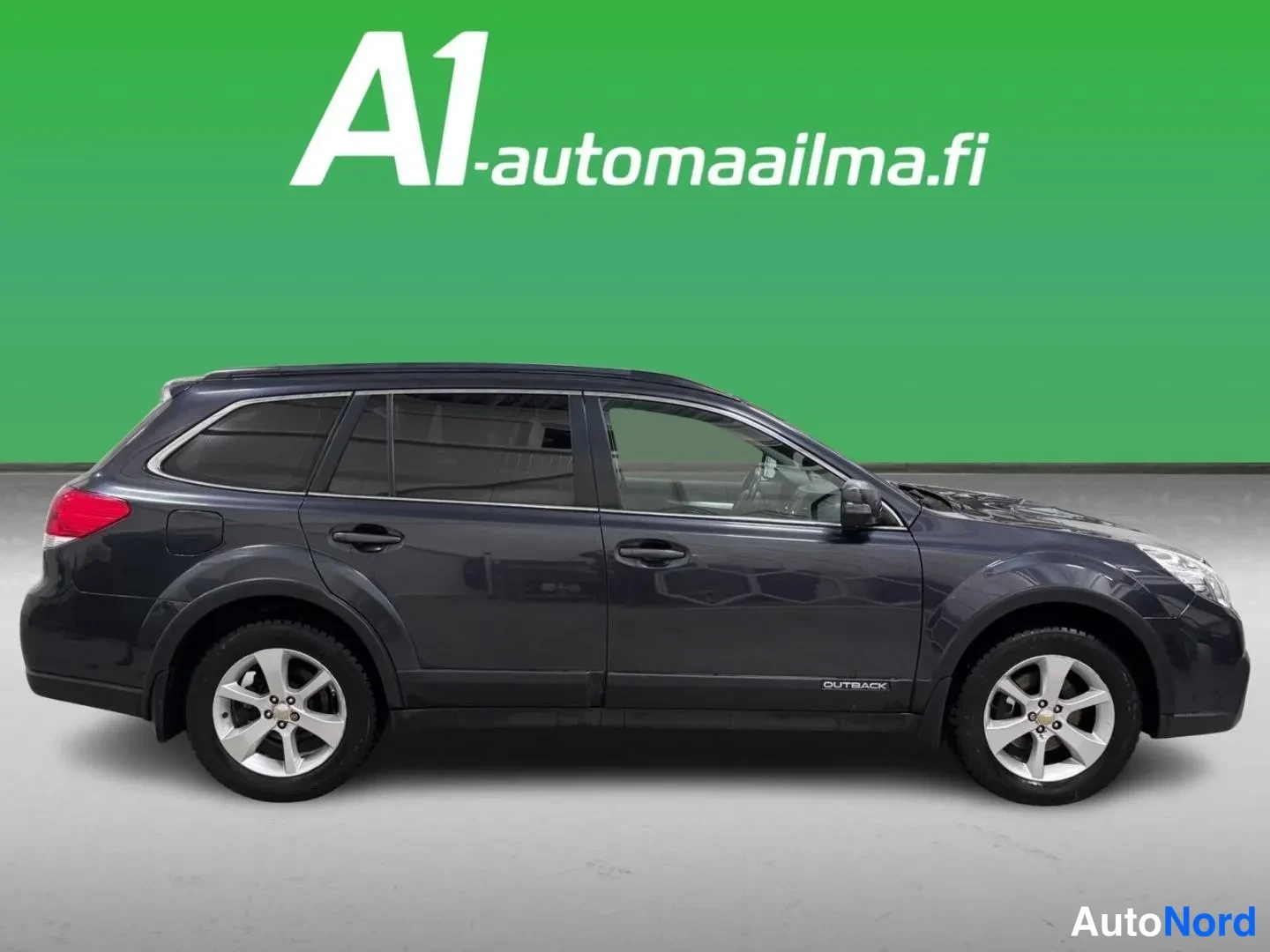 Etuviisto, nokka oikealle: Auton matkustajan puolen sivukulma: tummanharmaa farmariauto kokonaisuudessaan, keula kuvan oikeassa reunassa, hopeanväriset vanteet ja OUTBACK-merkintä