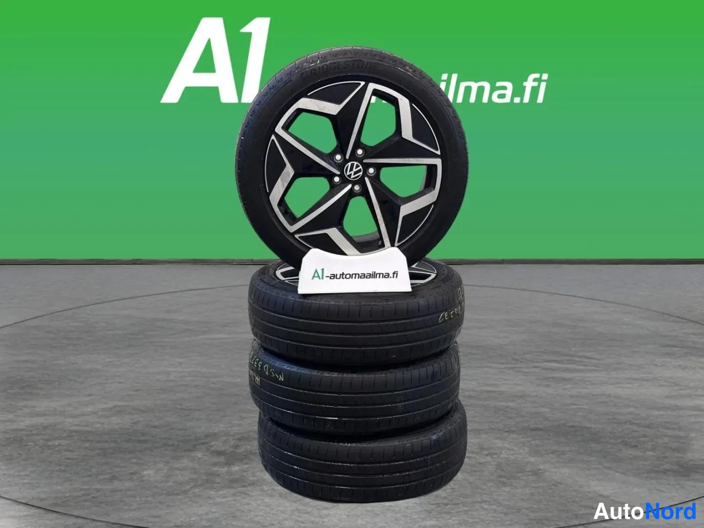 Yksityiskohta: Pino neljää Bridgestone-rengasta, päällimmäinen vasummalla VW-alumiinivanteella, A1-automaailman studio­taustaa vasten.