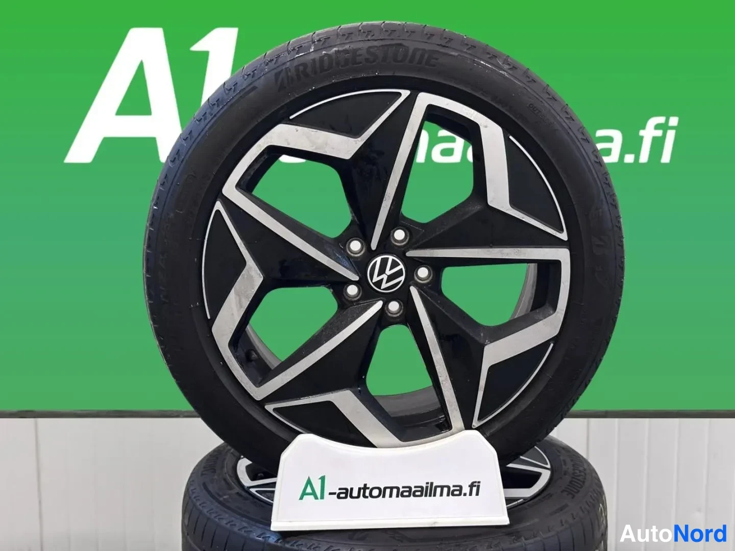 Yksityiskohta: Lähikuva musta-hopeasta viisipaltaisesta VW-alumiinivanteesta ja Bridgestone-renkaasta, A1-automaailman logotaustaa vasten.