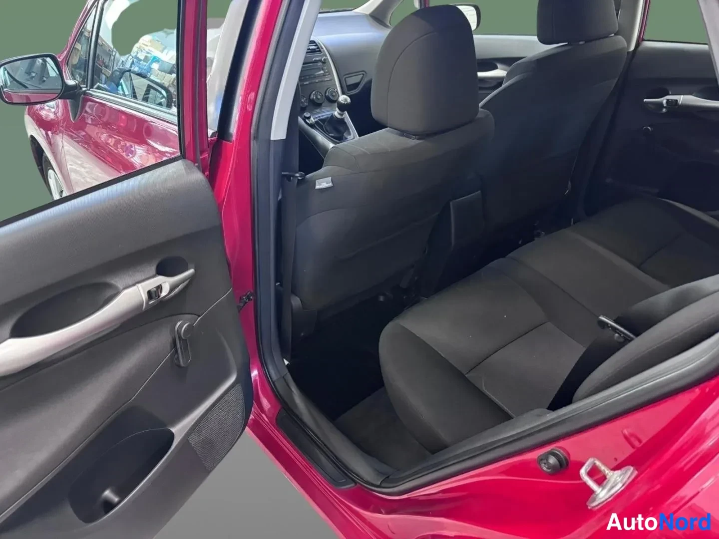 Yksityiskohta: Punaisen Toyota Auriksen takaistuin ja takaosan lattia avatun takaovan läpi; kaksiosainen takaistuin ja lattiamatto näkyvät, yläosissa valaistus ja kattokahvat.