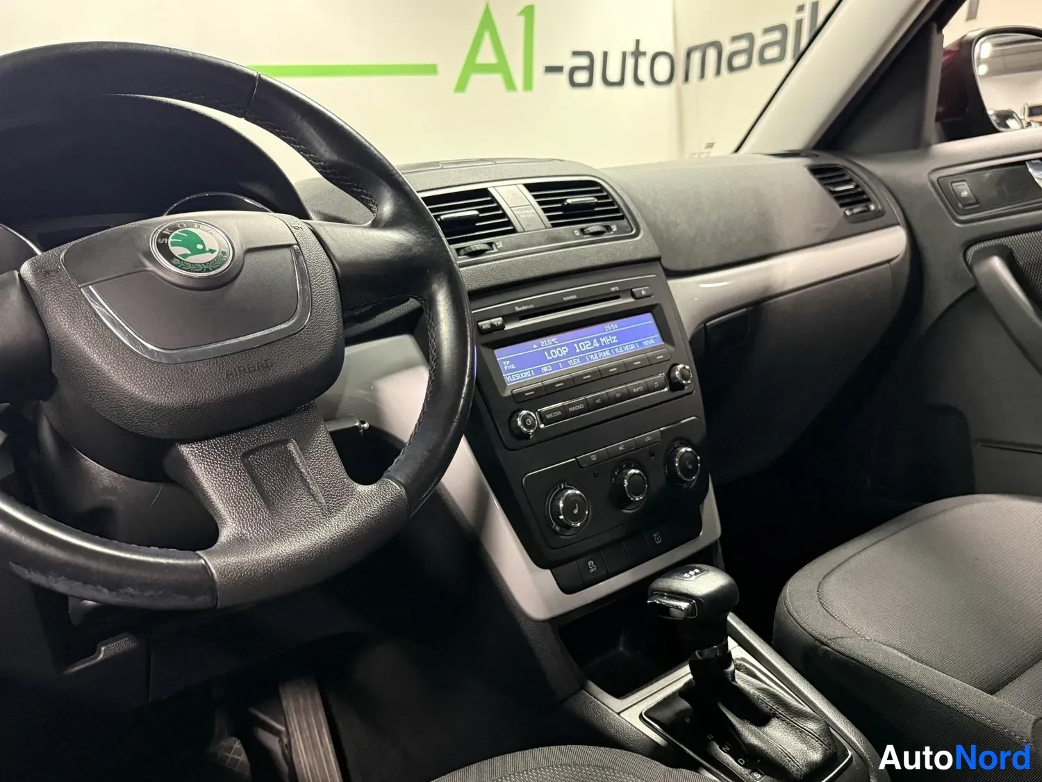 Yksityiskohta: Ohjauspylan alue lähikuvassa: Skoda-merkillä varustettu ohjauspyörä, radio- ja ilmastointilaitteet näytöllä sekä automaattivaihteen valitsin.