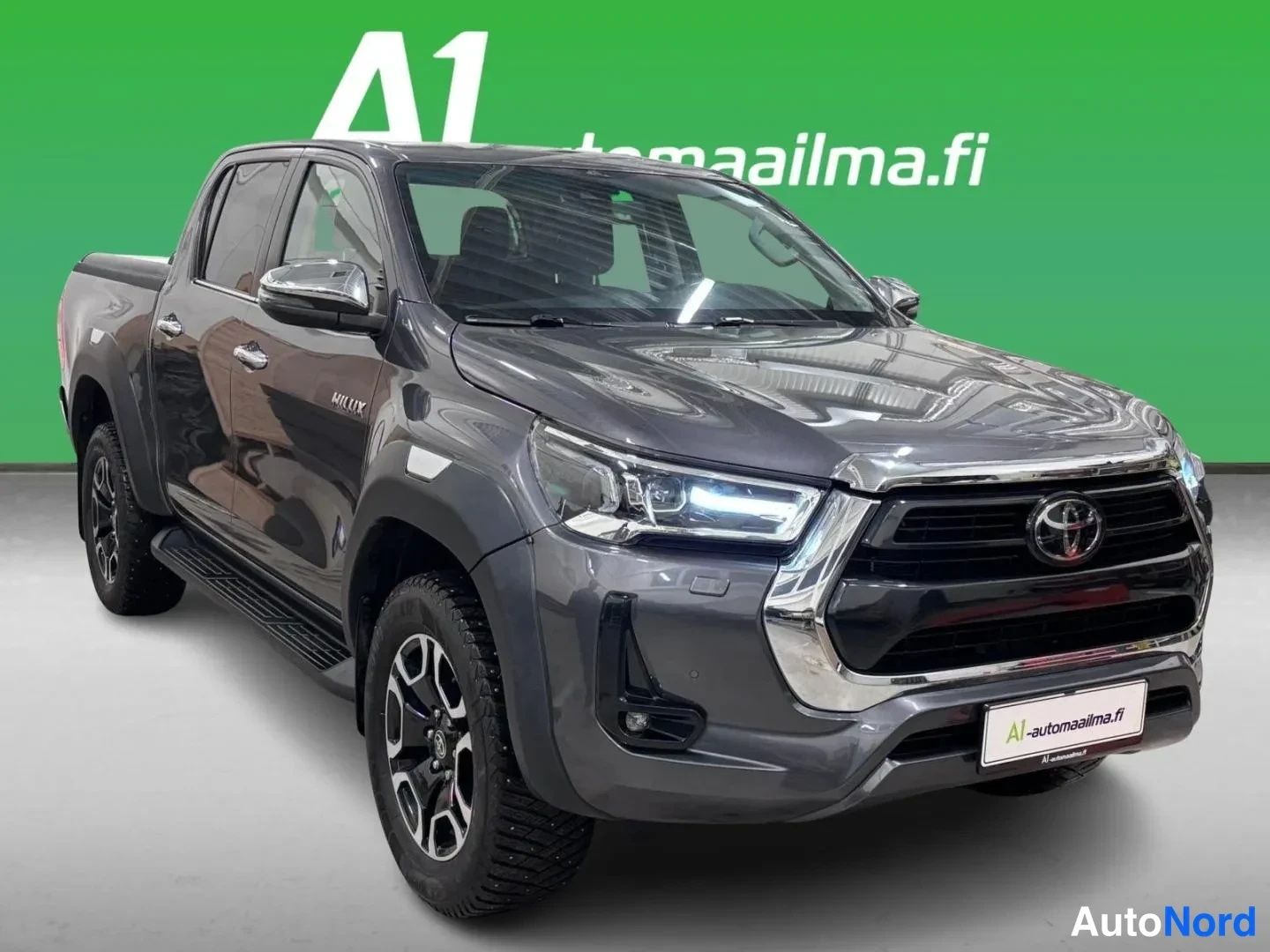 Etuviisto, nokka oikealle: Tummanharmaa Toyota Hilux -laccapickup etukulmasta, keula oikealla, kylki ja kuormasänky näkyvät.