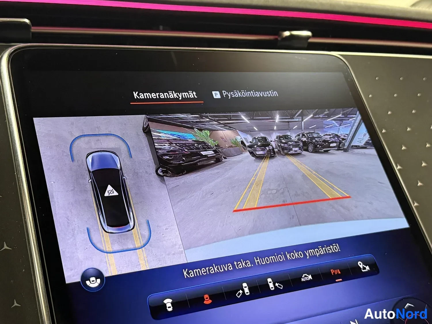 Muu: Auton infotainment-näytöllä näkyy kamerafunktioiden valikko otsikolla 'Kameranäykymät' sekä pysäköintikameran kuva pysäköintialueelta apuviivoineen. Ruudun alao