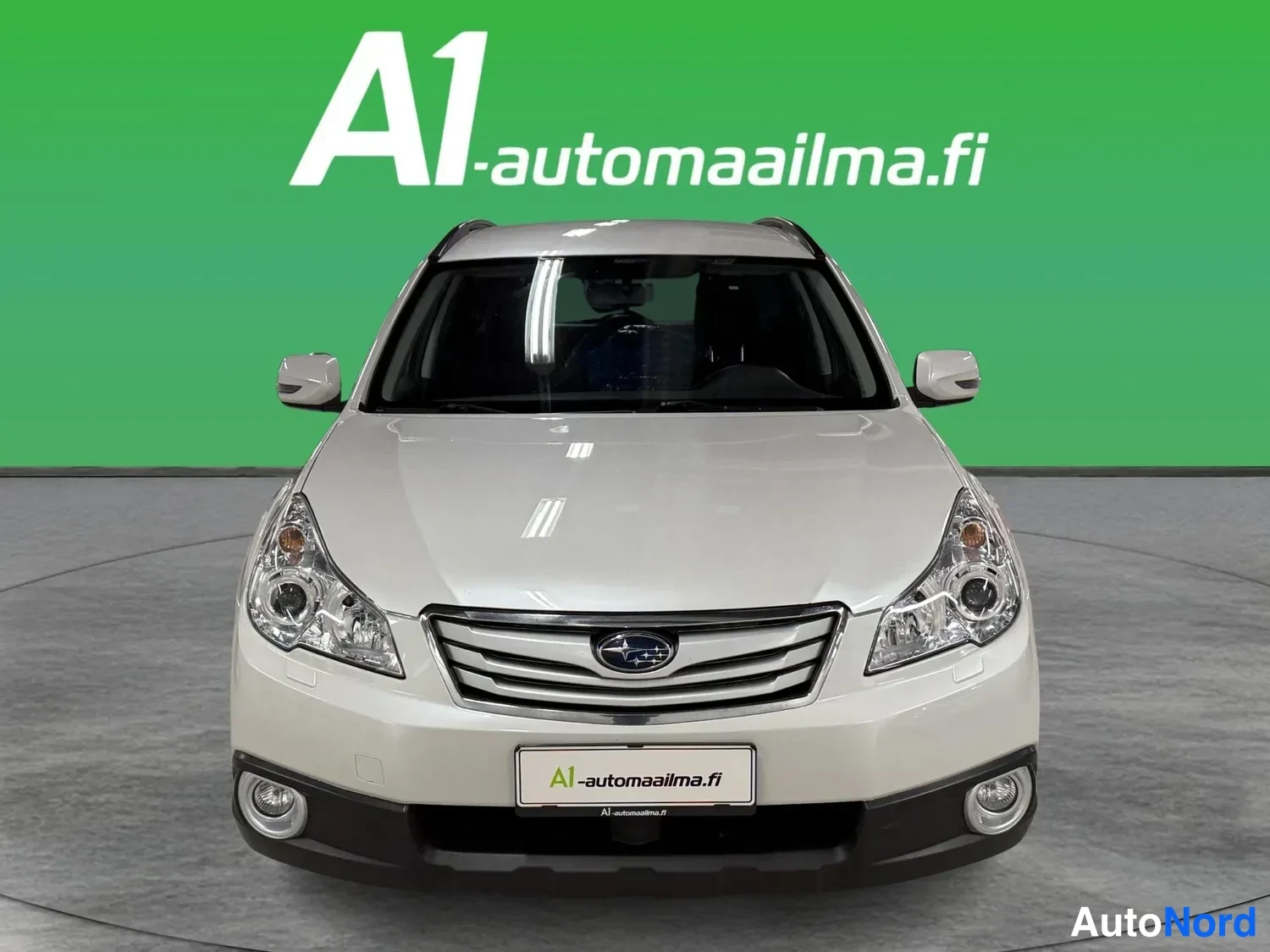 Edestä: Subaru Outback -farmari suoraan edestäpäin valokuvattu, valkoinen kori, etupuskurissa A1-automaailma.fi -kyltti, studiotrkTausta.
