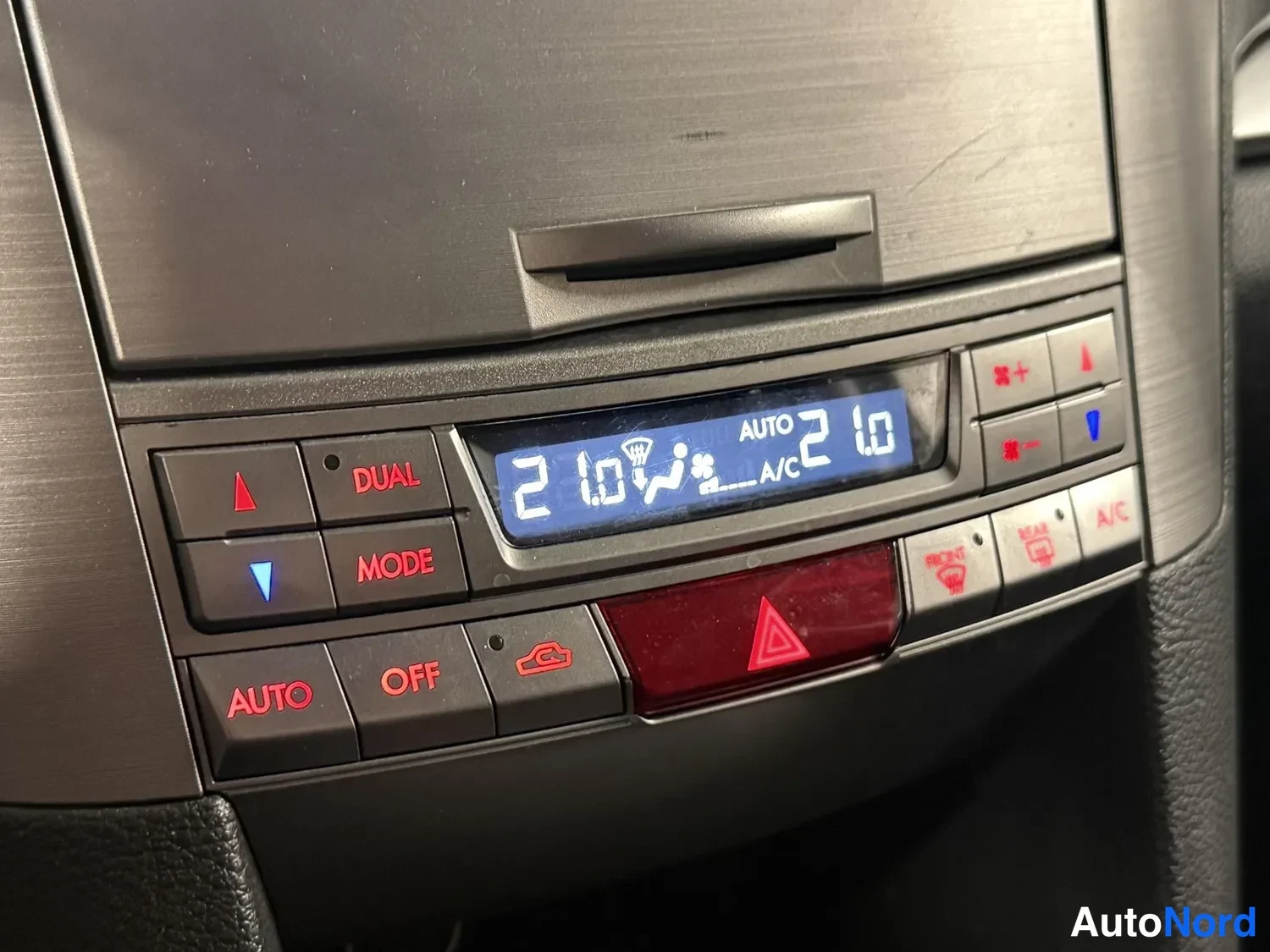 Yksityiskohta: Lähikuva auton ilmastoinnin ohjauspaneelista: näytössä AUTO ja A/C -merkit sekä lämpötilat 2 ja 1.0, painikkeet DUAL, MODE, AUTO ja OFF punaisin tekstein.