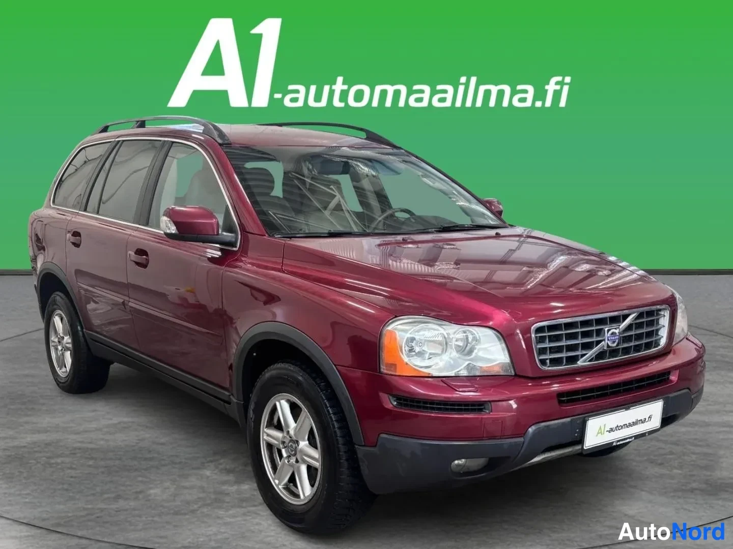 Etuviisto, nokka oikealle: Tummanpunainen Volvo XC90 kuvattu edestä oikealta kolmeneljäsosakulmasta; konepelti, etugrigaatti ja kylki näkyvät vihreää taustaa vasten.