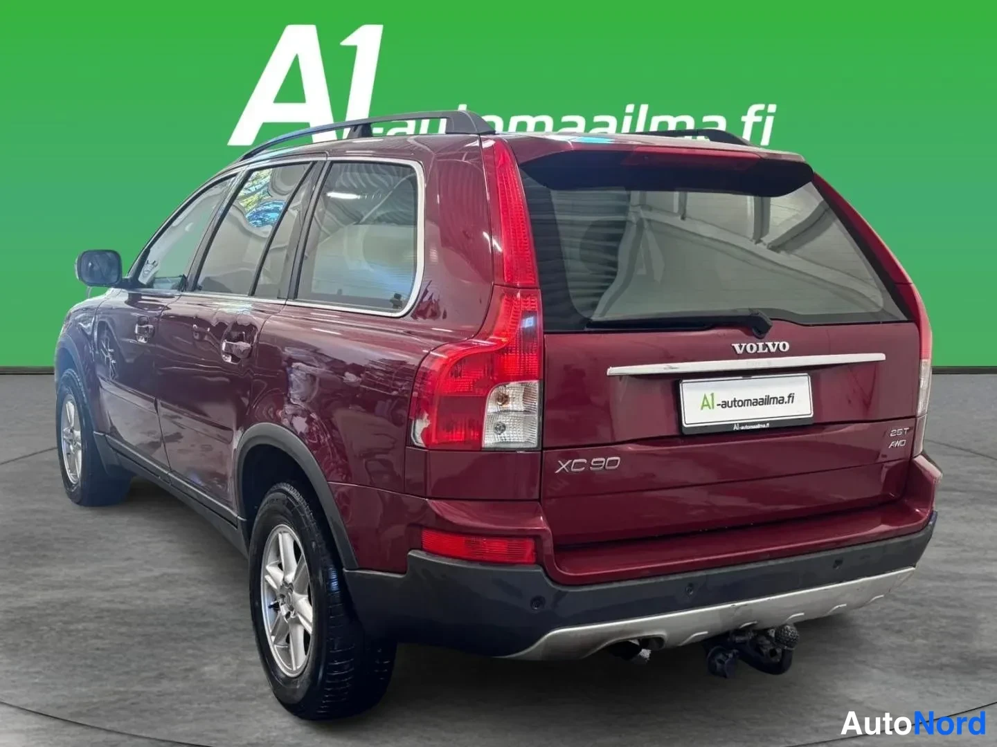Takaviisto: Tummanpunaisen Volvon takanäkymä vasemmalta kolmeneljäsosakulmasta: XC90-merkki, VOLVO-teksti, takavalot ja takapuskuri näkyvät vihreää taustaa vasten.