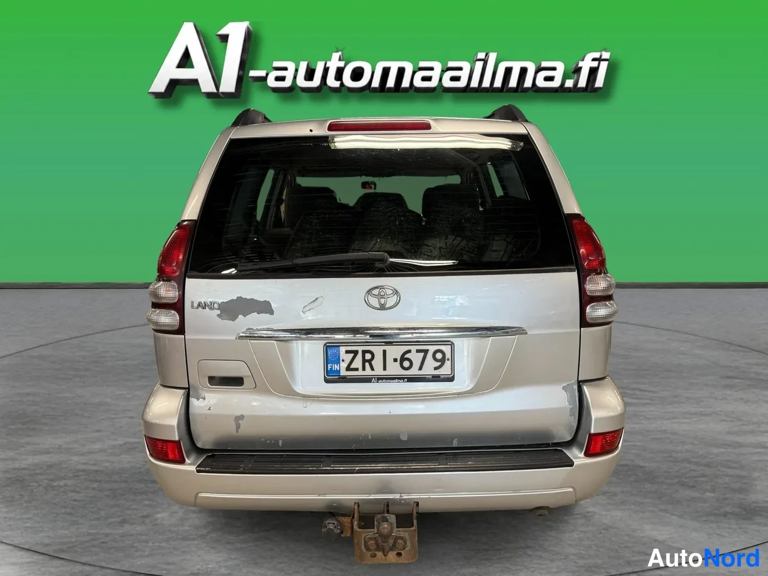 Takaa: Auton takaosa suoraan takaa: hopea Toyota, rekisteritunnus, vedokoukku ja Logo-merkintä takapuskurissa, takaluukussa Land Cruiser -teksti.