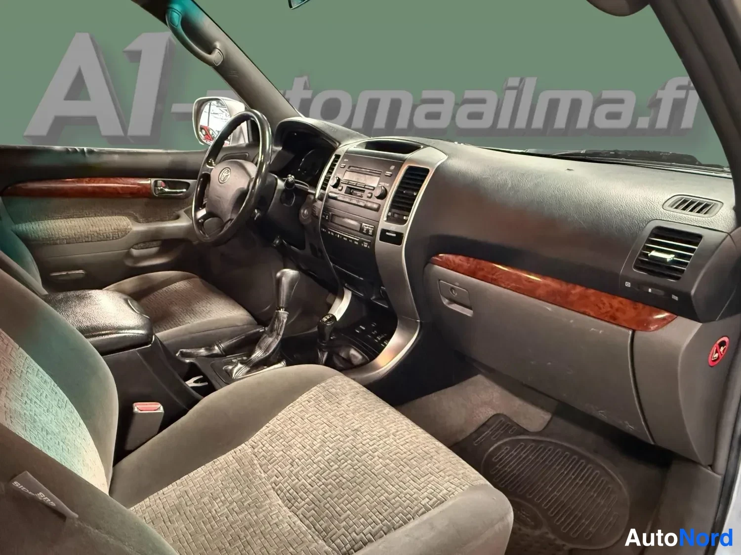 Edestä: Ohjaamo vasemmalta takaa kuvattuna: kuljettajan istuin ja oviverhoilu, Toyota-ohjauspyörä, keskikonsolin radio ja vaihdevipu, hanskalokero sekä puukoristeinen k