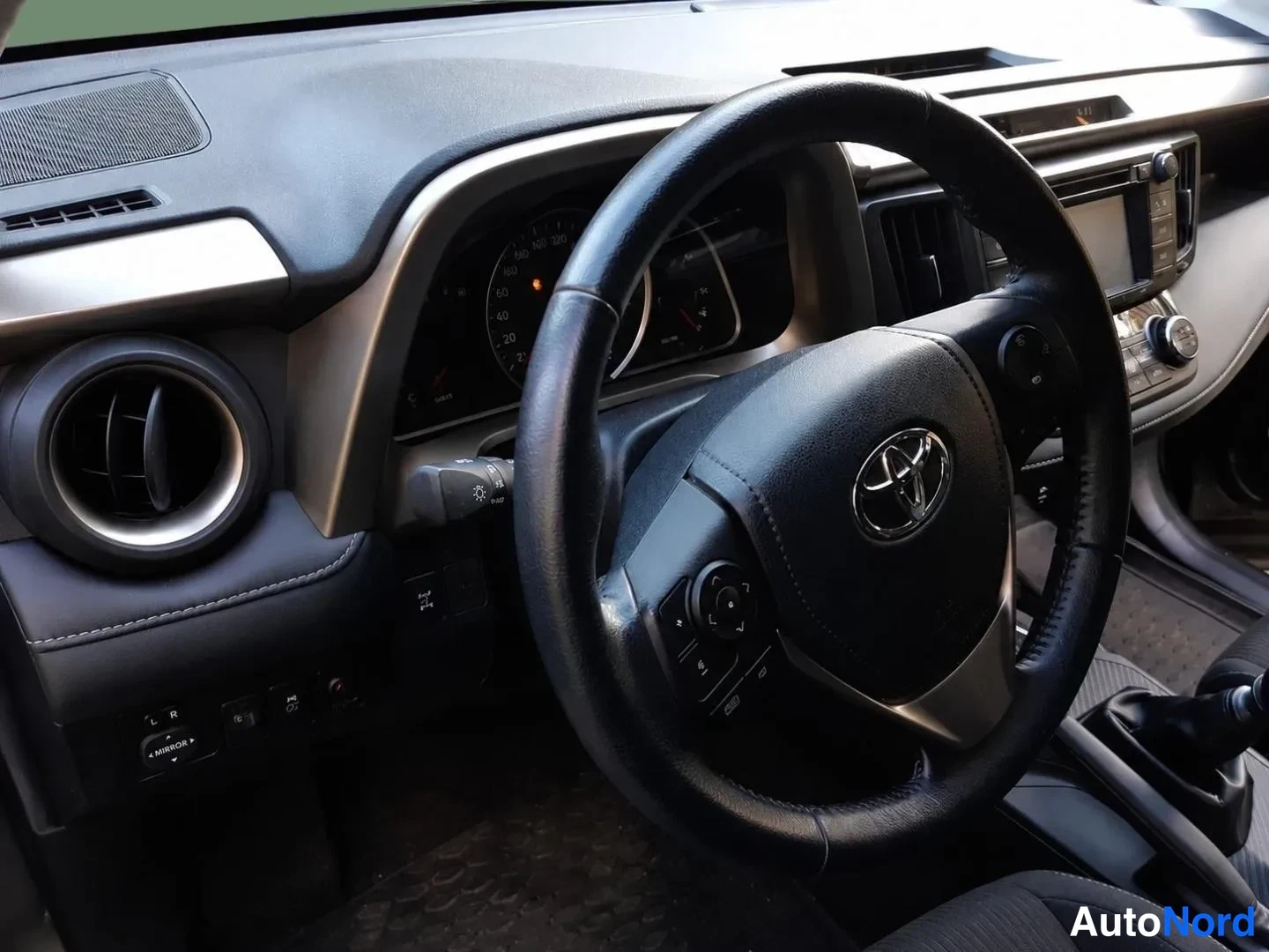 Sivulta: Ohjaamon näkymä: Toyota-merkkinen ohjauspyörä, mittaristo ja tumma kojelauta.