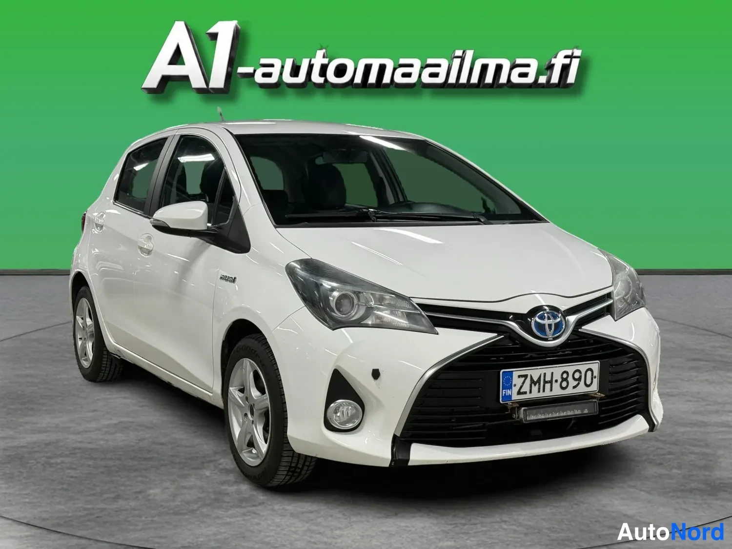 Etuviisto, nokka oikealle: Valkoinen Toyota Yaris -henkilöauto etuviistossa vasemmalta, etupanssari ja rekisterikilpi näkyvissä.
