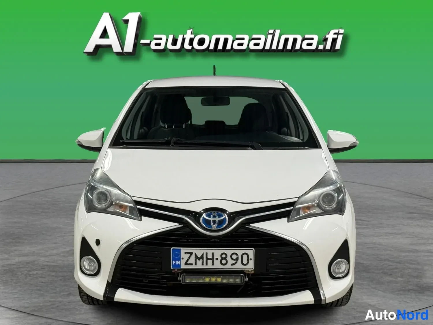 Edestä: Valkoinen Toyota Yaris suoraan edestä, Toyota-kehtokeula ja rekisterikilpi näkyvissä.