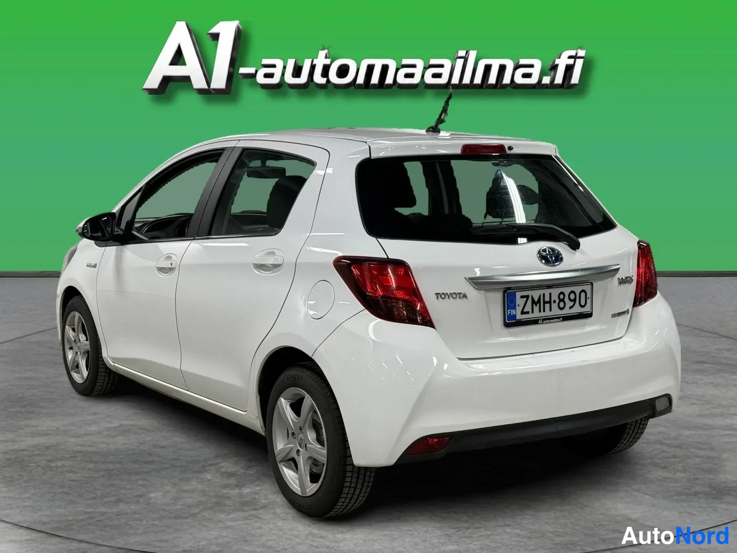 Takaviisto: Valkoinen Toyota Yaris takaviistosta vasemmalta, takavalot, Toyota-merkki ja rekisterikilpi näkyvissä.
