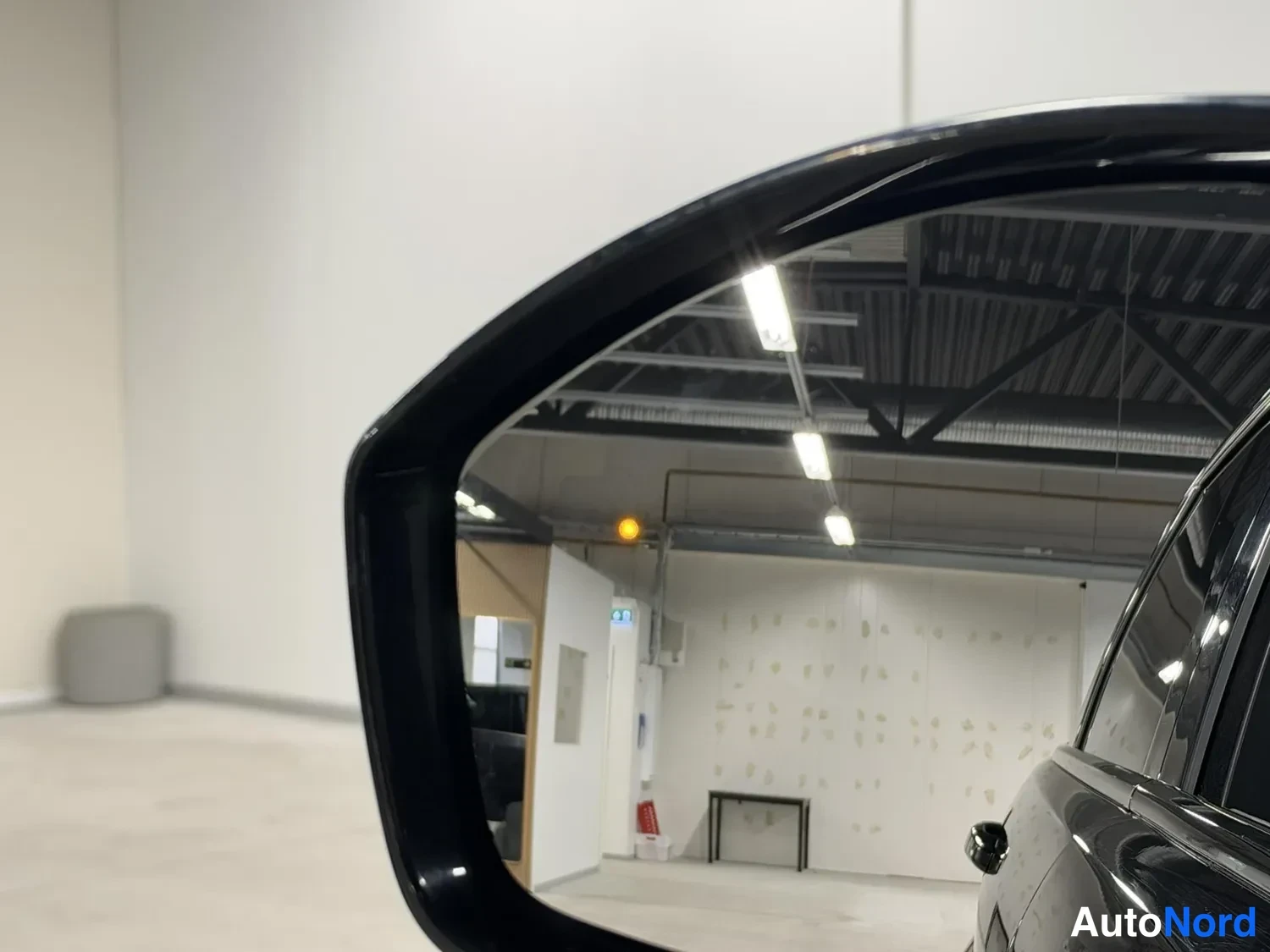 Yksityiskohta: Takapeilin lähikuva mustasta autosta sisätiloissa; peilin kuvassa halli, valaisimet ja ovipuoli.