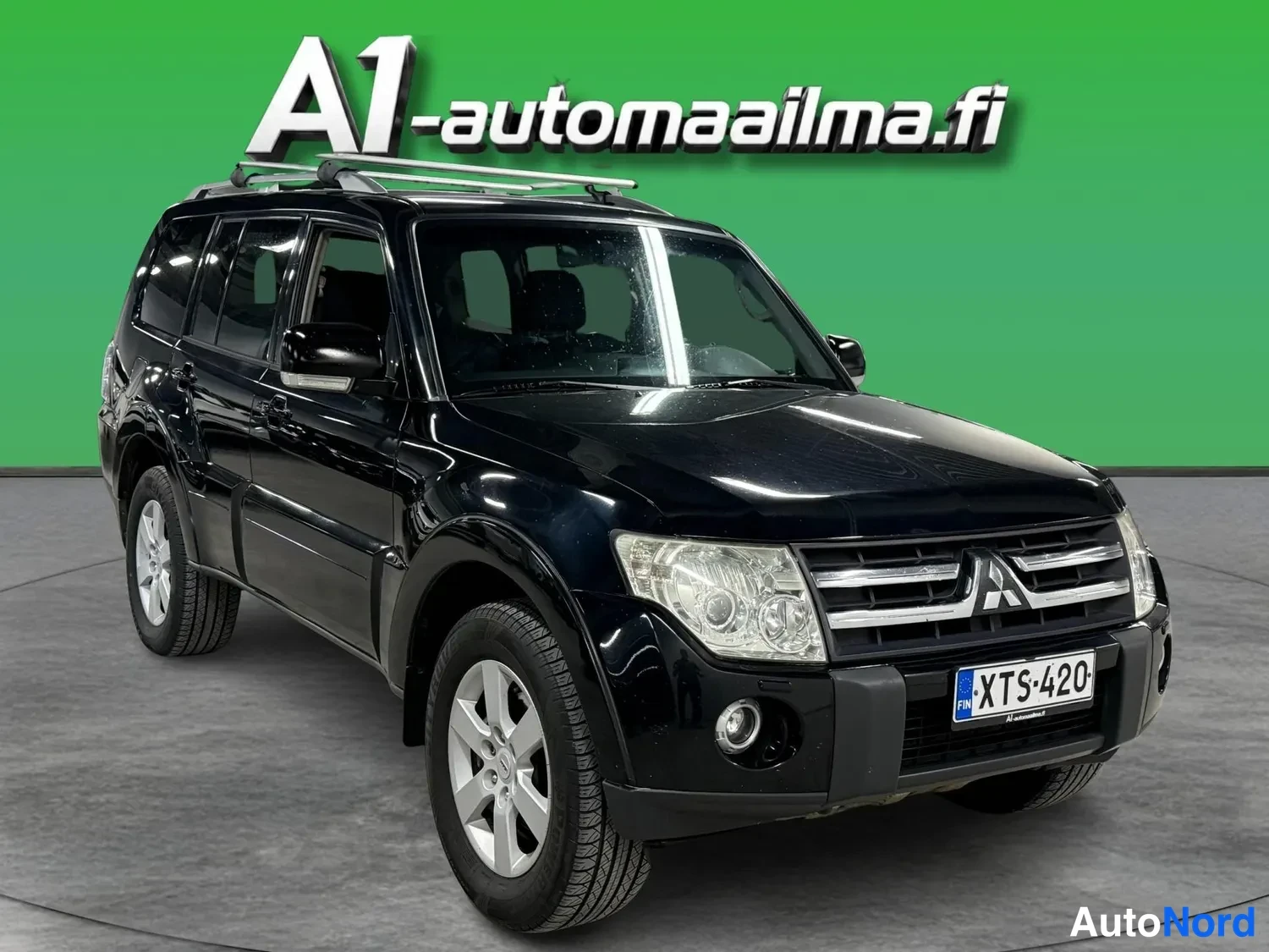 Etuviisto, nokka oikealle: Musta Mitsubishi Pajero -maasturi kolmivartalokulmassa edestä oikealta, rekisteritunnus, kattotelineet ja hopeiset kevytmetallivanteet.