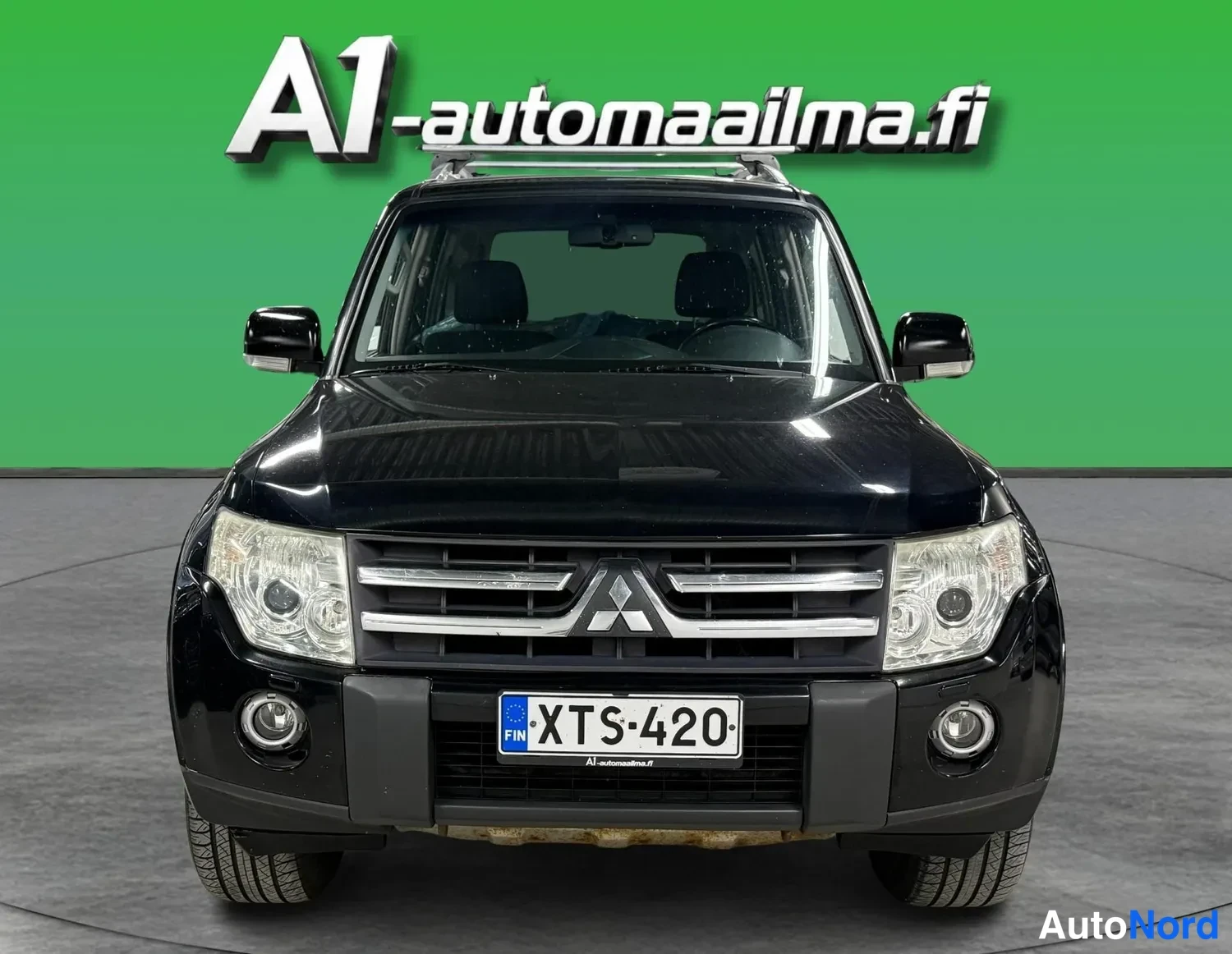 Edestä: Musta Mitsubishi Pajero suoraan edestä, rekisteritunnus, Mitsubishi-tunnus etusäleikössä ja kattotelineet.