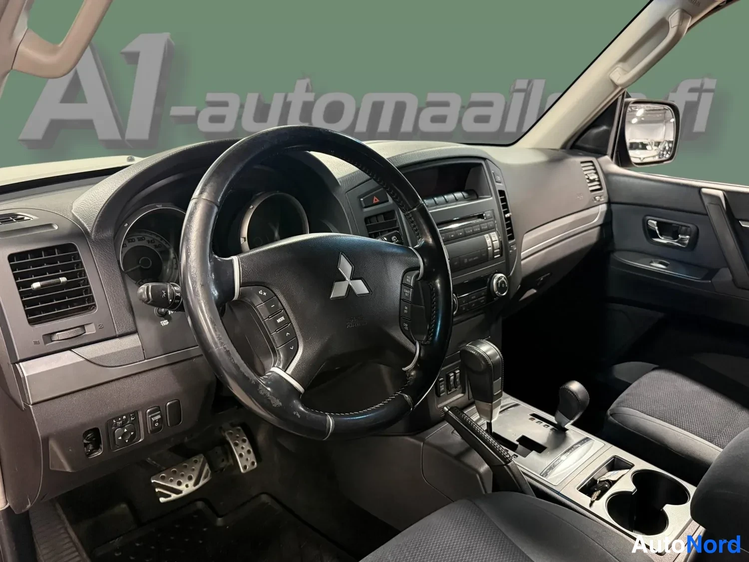 Muu: Ohjaamon yleiskuva: Mitsubishi-merkillä varustettu ohjauspyörä, kojelauta, radio ja automaattivaihteen valitsin; vasen etuovi auki.