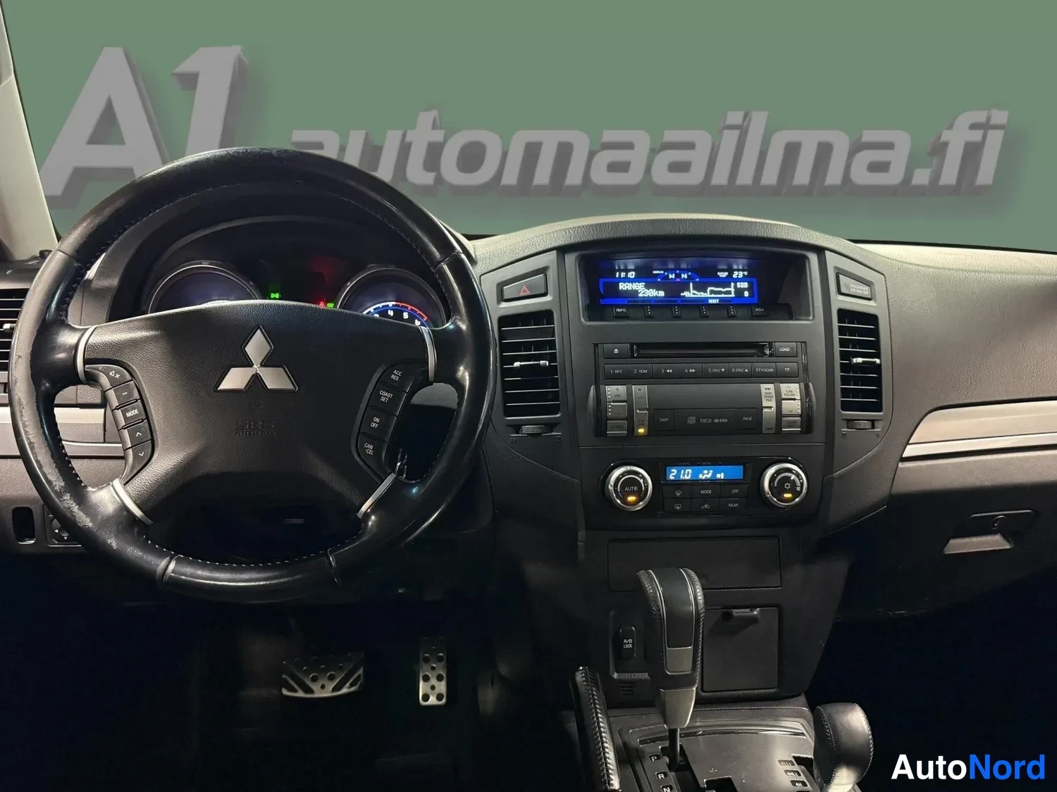 Edestä: Mitsubishi-merkkinen ohjauspyörä ja tumma kojelauta: radiota, infonäyttö jossa teksti 'RANGE 288km' sekä kaksivyöhykeilmastointi.