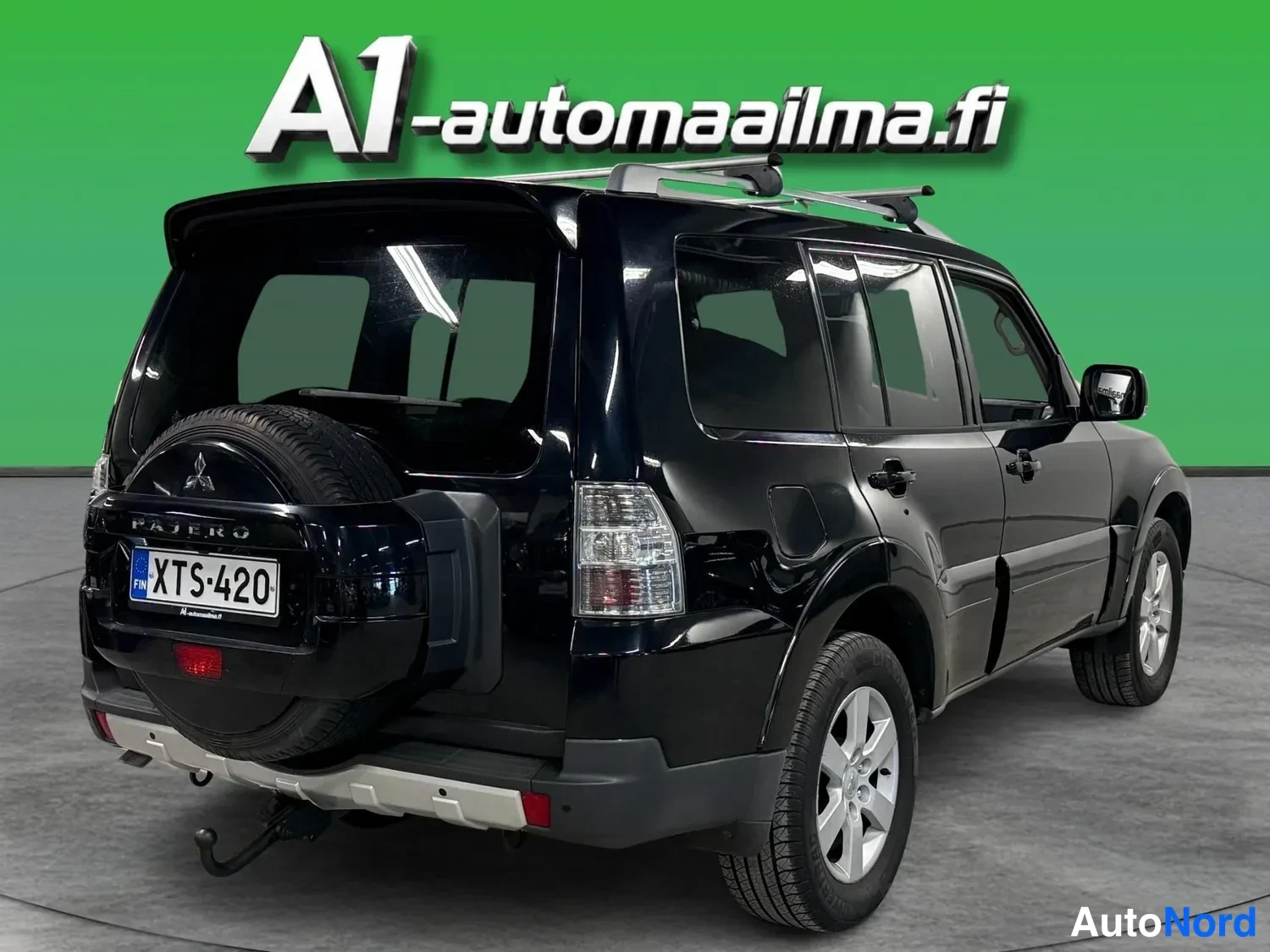 Takaviisto: Mustan Mitsubishi Pajeron takaosa ja vasen sivuprofiili kolmasosa-kulmasta; kattoteline, varapyörä ja vetokoukku näkyvissä.