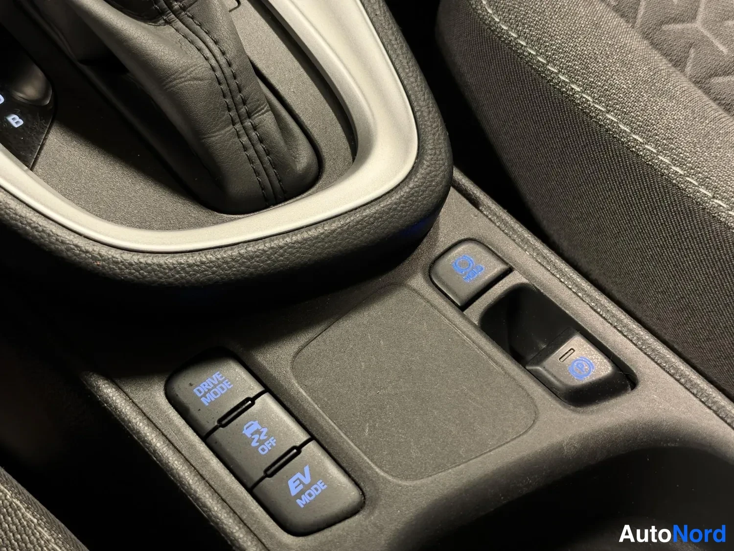 Yksityiskohta: Keskikonsolin ajotilanapit lähikuvassa: DRIVE MODE, TRC OFF ja EV MODE -napit sekä elektroniinen parkkirijätjöt; oikealla istuinverhoilu.