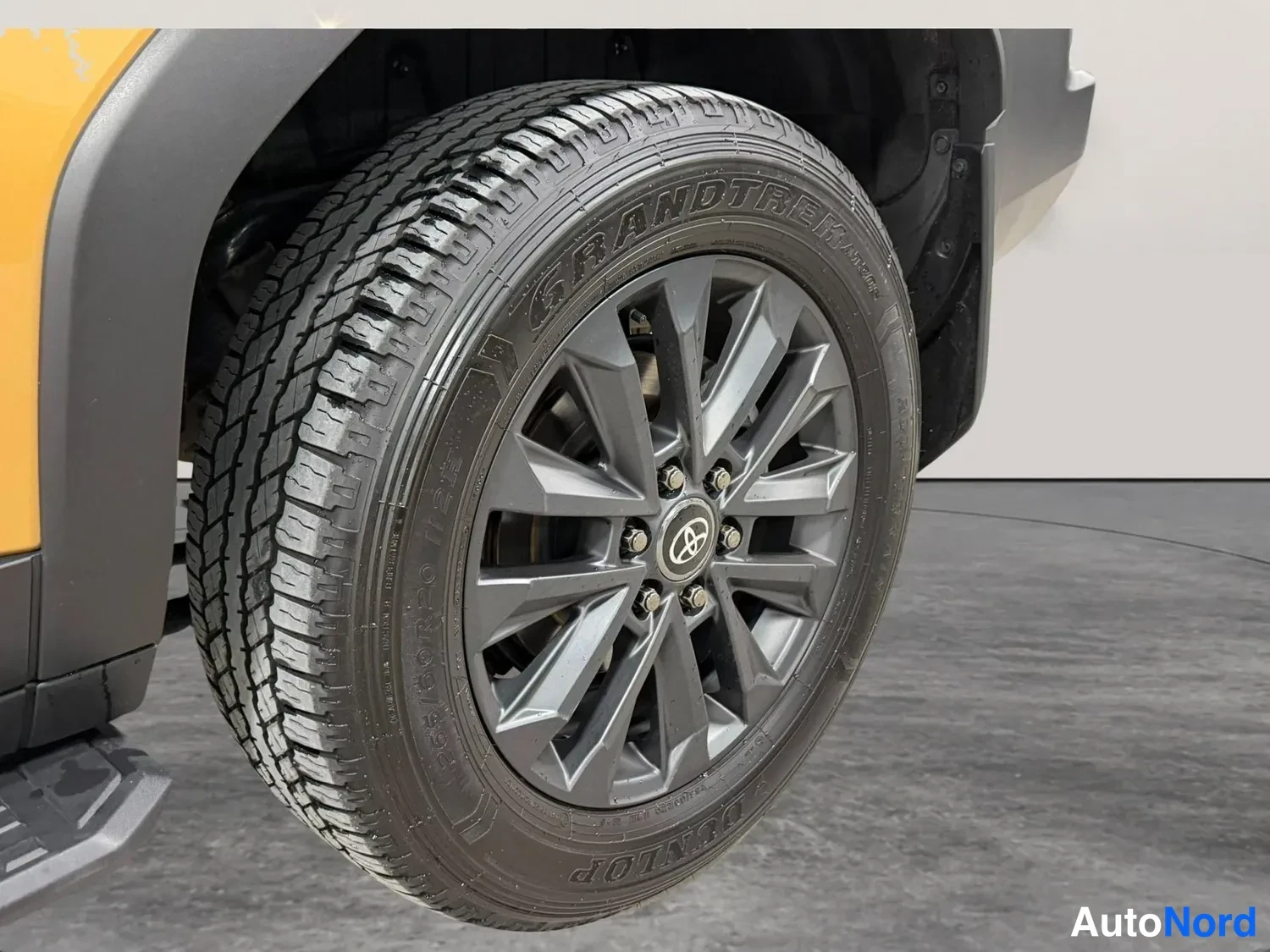 Muu: Lähikuva auton pyörästä: harmaa kevytmetallivanne Toyota-emblemillä ja Dunlop Grandtrek -keskareeri.
