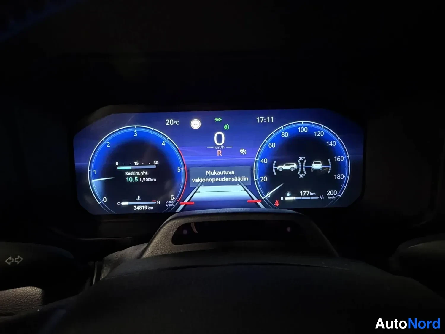 Muu: Autojen kojelaudan mittaristö näytön keskellä, nopeus 0 km/h ja vaihde R, kilometrilukema 34819 km näkyvillä.