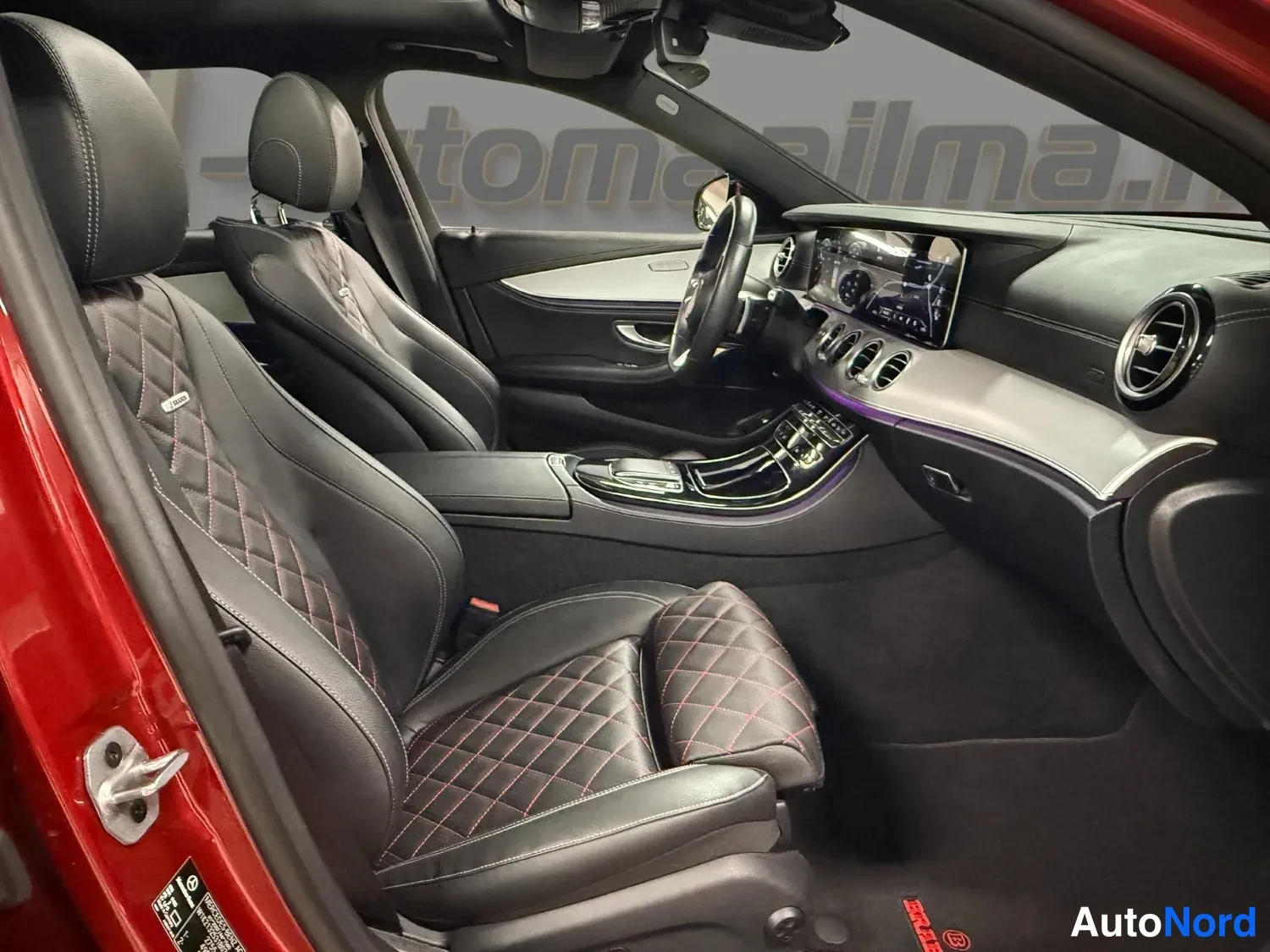 Muu: Avoimen etuovin kuvattu punaisen auton sisusta: kaksi mustaa kiiltavanahkista etuintakaa punaisilla ristipistoilla, mittaristo ja keskinaytto, ohjauspyora ja ke