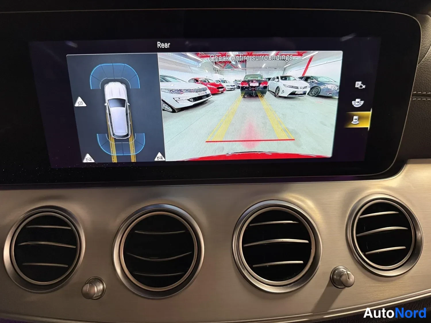 Muu: Infotainment-näyttö näyttää peruutuskamerakuvaa tekstillä 'Rear' ja 'Check entire surroundings' sekä kaavion auton sijoittumisesta, alla ilmastointisuuttimet.